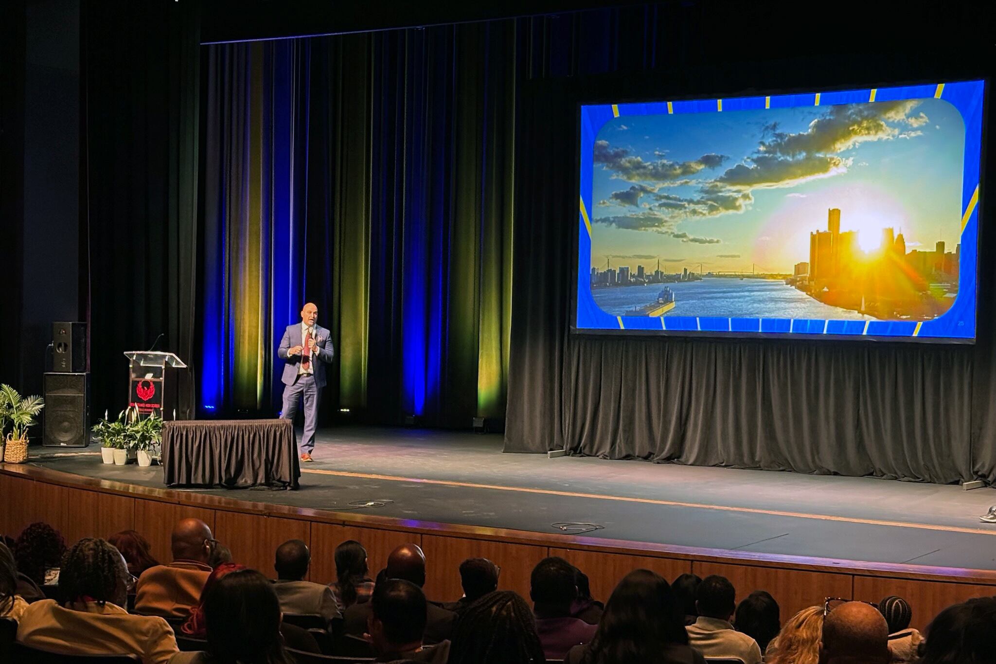 A man in a suit stands on stage with people sitting in rows of chairs in the foreground.