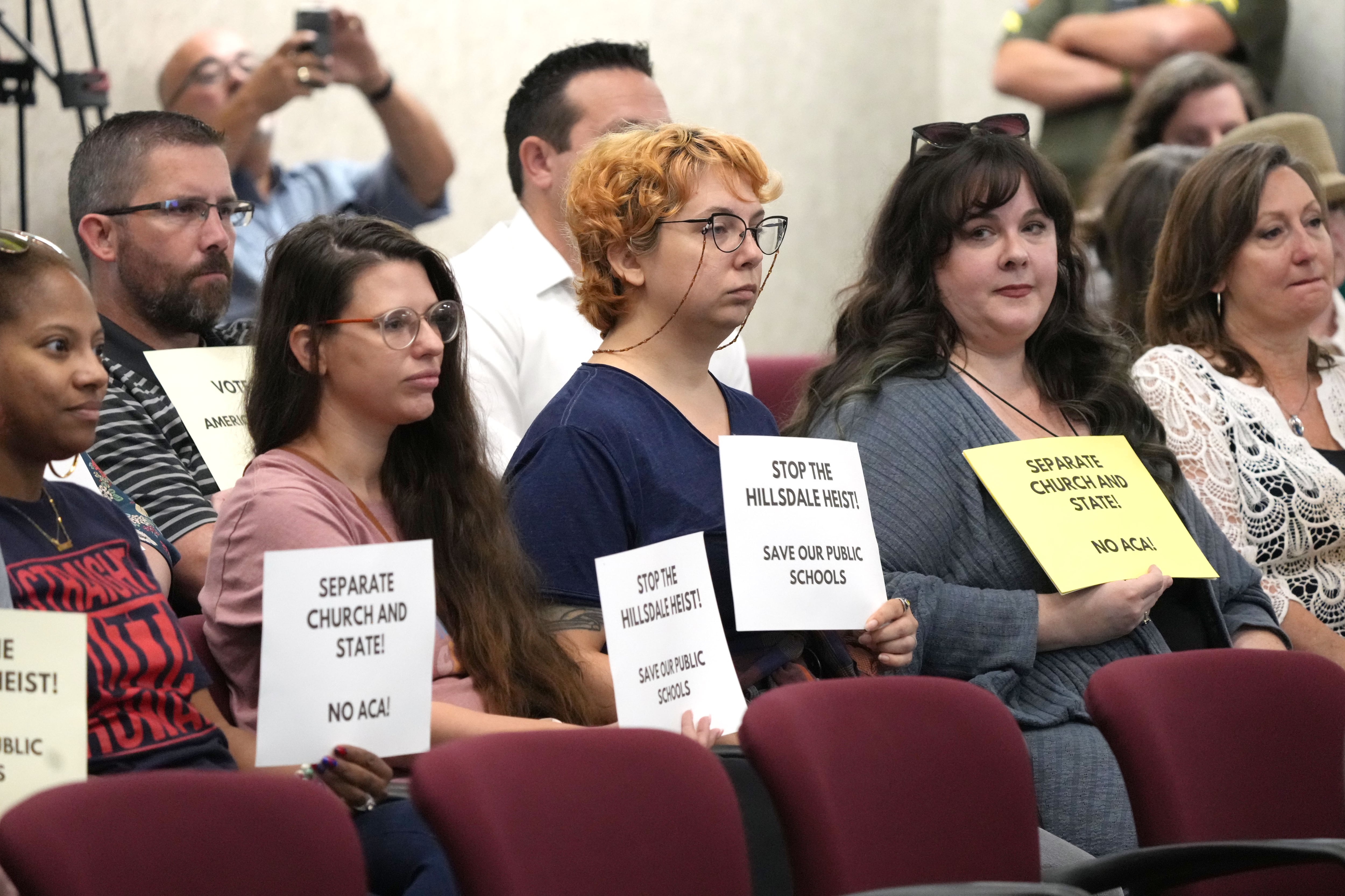Five women seated during a meeting hold signs opposing Hillsdale College.