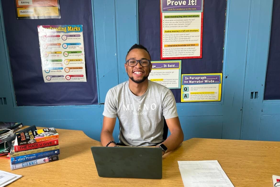A teacherwearing a grey shirt sits in his classroom. He is at his computer.