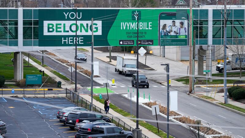 A green sign on a building’s connecting bridge reads “YOU BELONG HERE”, and advertises Indiana’s Ivy Tech Community College. Under the bridge, cars drive down a street near a college campus.