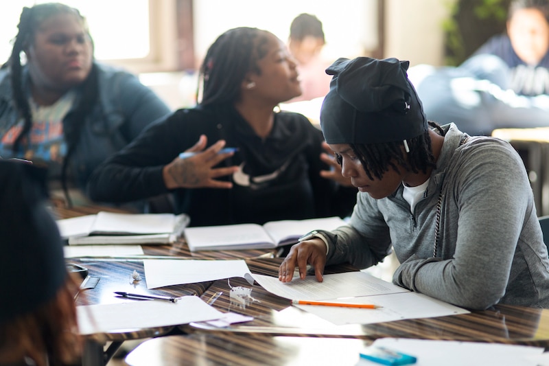 High school students work on assignments while sitting at desks in a classroom.