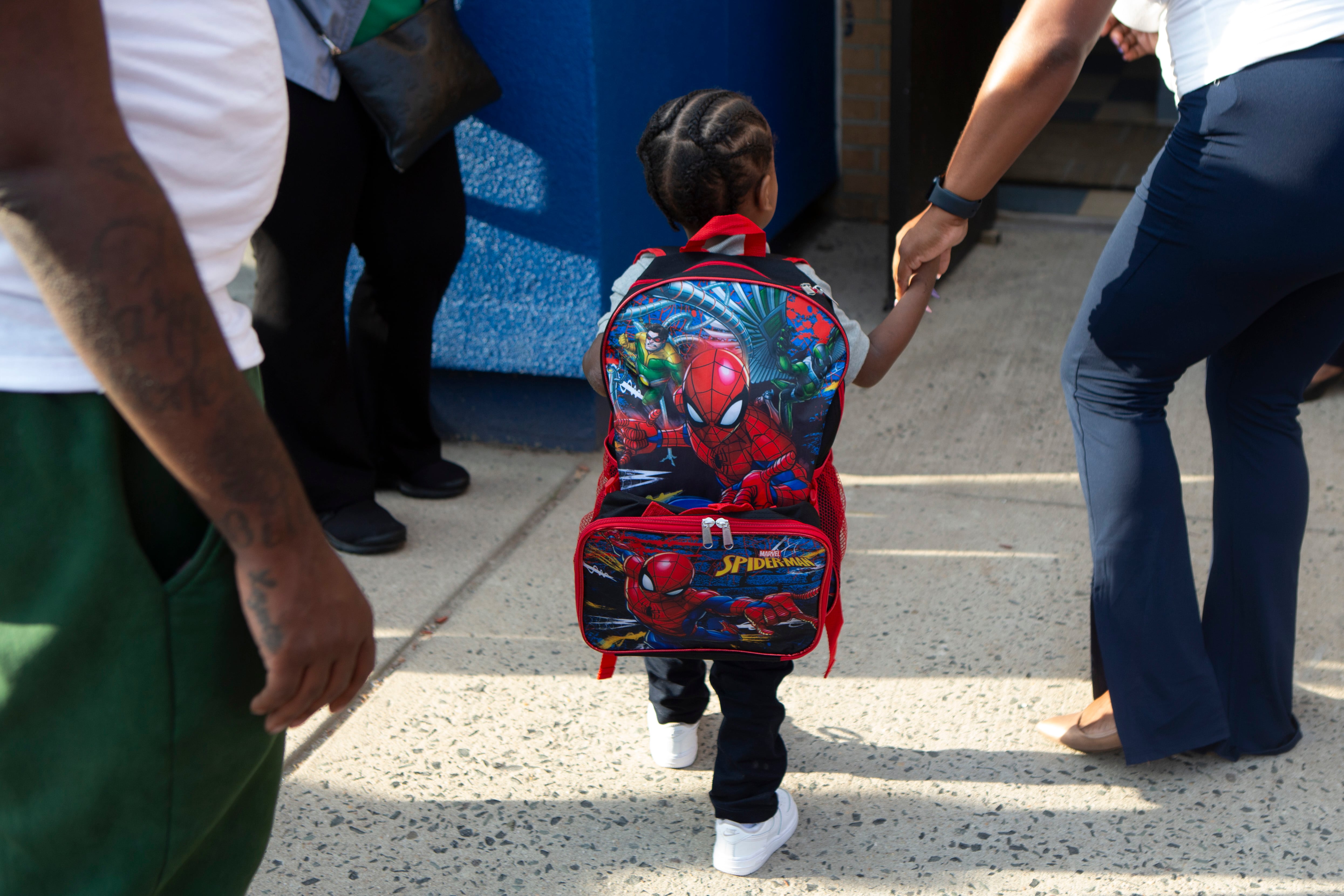 A young student with braids and wearing a large red Spiderman backpack, walks towards the school building holding the hand of an adult.