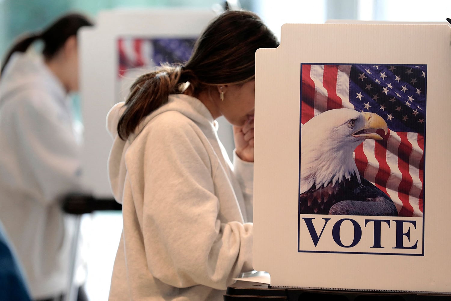 A young person with long brown hair and wearing a white sweater is voting inside of a voting cubicle with an American flag and an eagle with the word "VOTE," on the side.