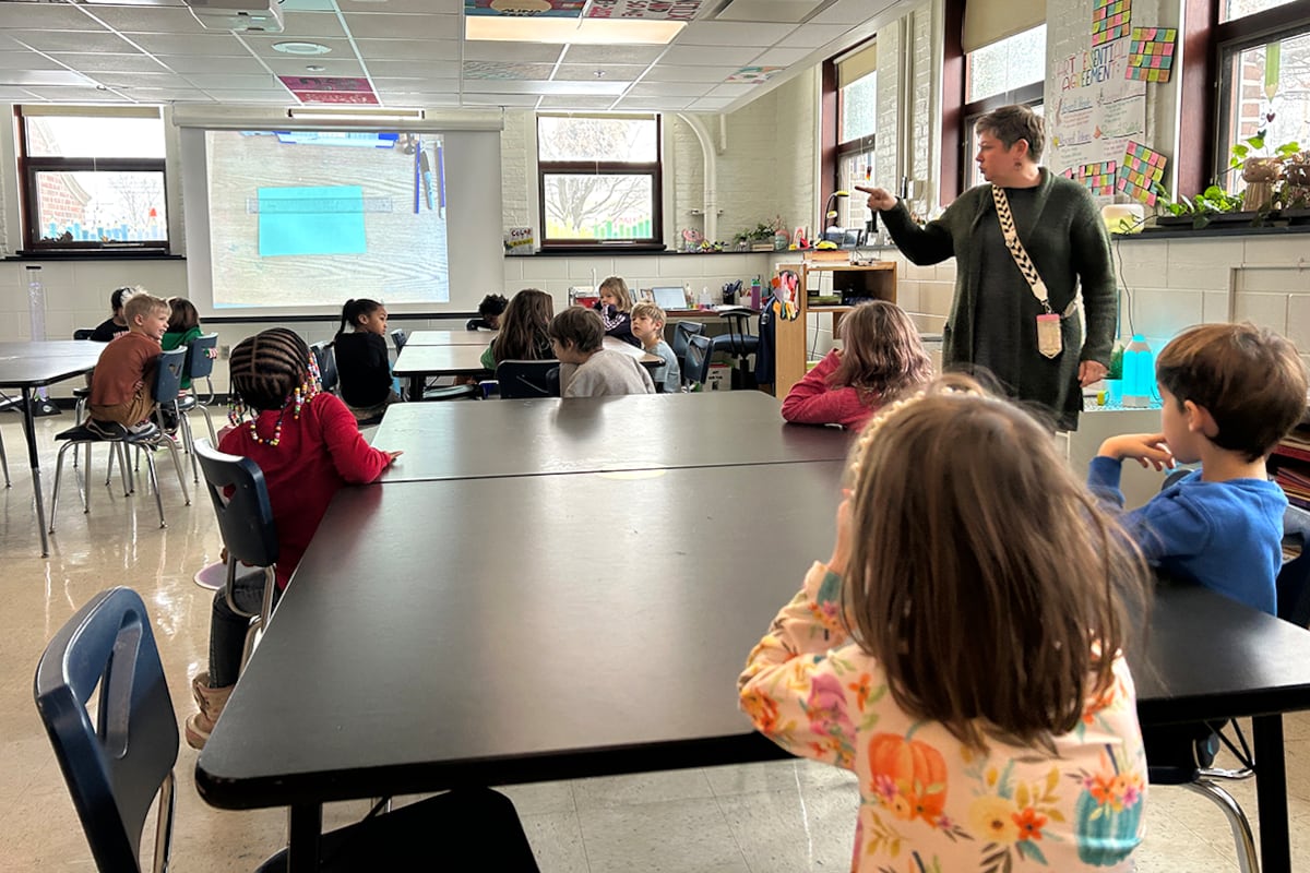 Students sit at a table while a teacher stands to the right.
