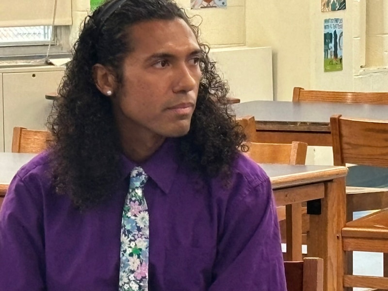A man with long dark hair and wearing a purple dress shirt and a colorful tie sits in a classroom.