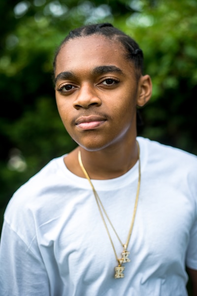 A portrait of King Bethel, who is wearing a white shirt and two gold necklaces with the letter K on them.