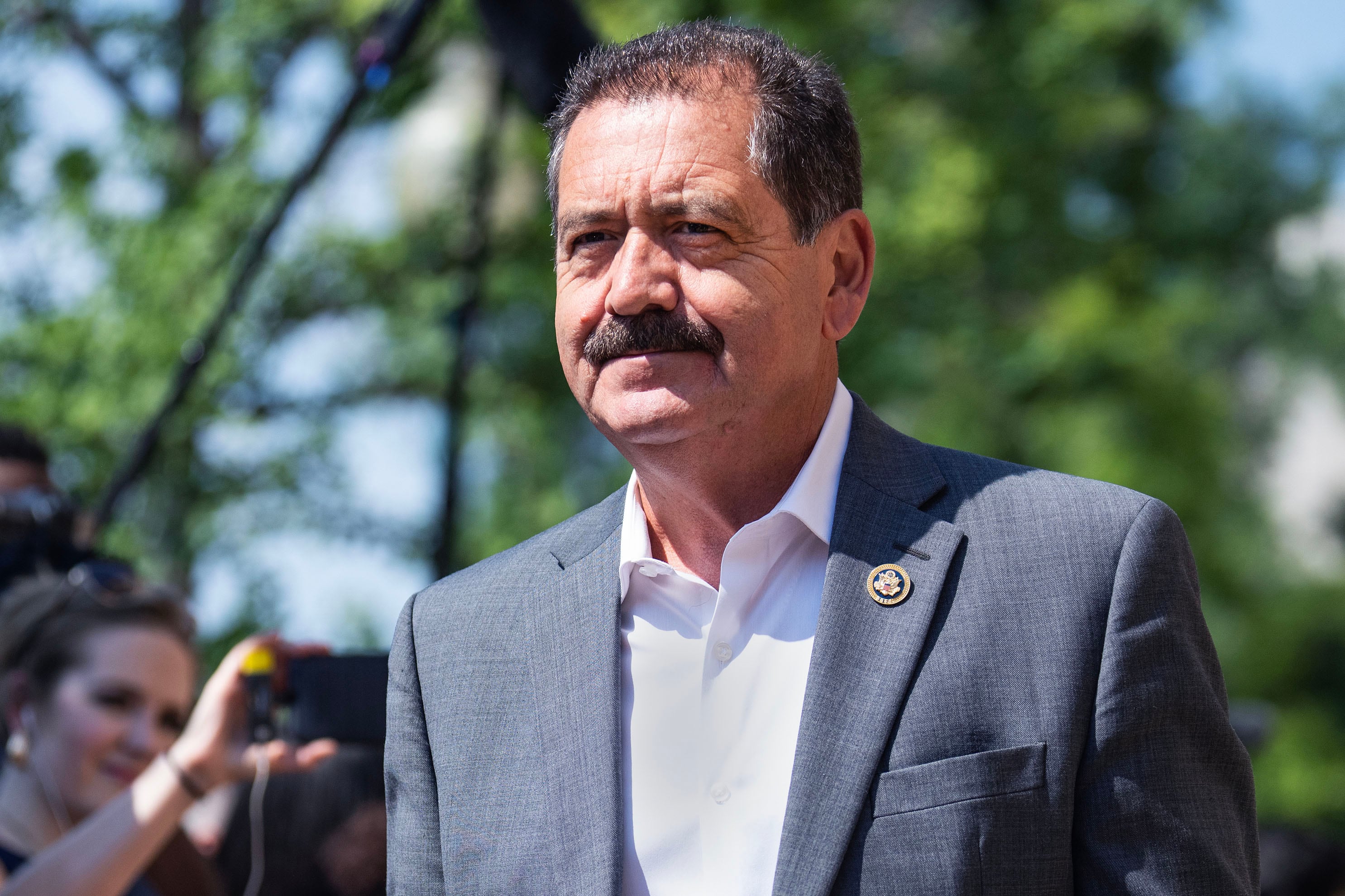 A man in a suit stands outside in the sun with green trees in the background.
