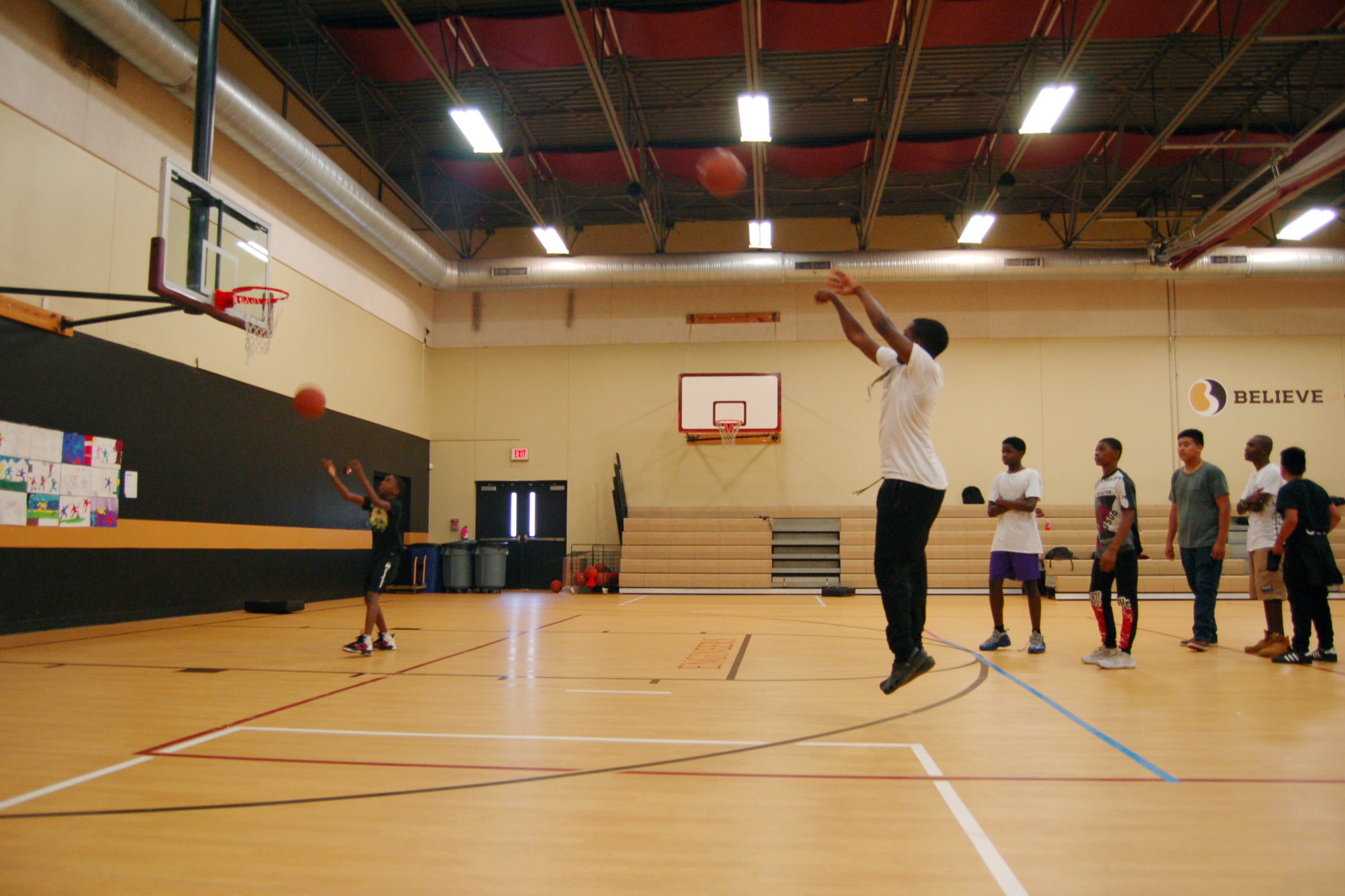 A boy jumps, still in the air, throwing a basketball toward the net. A line of students wait behind him in a gymnasium.