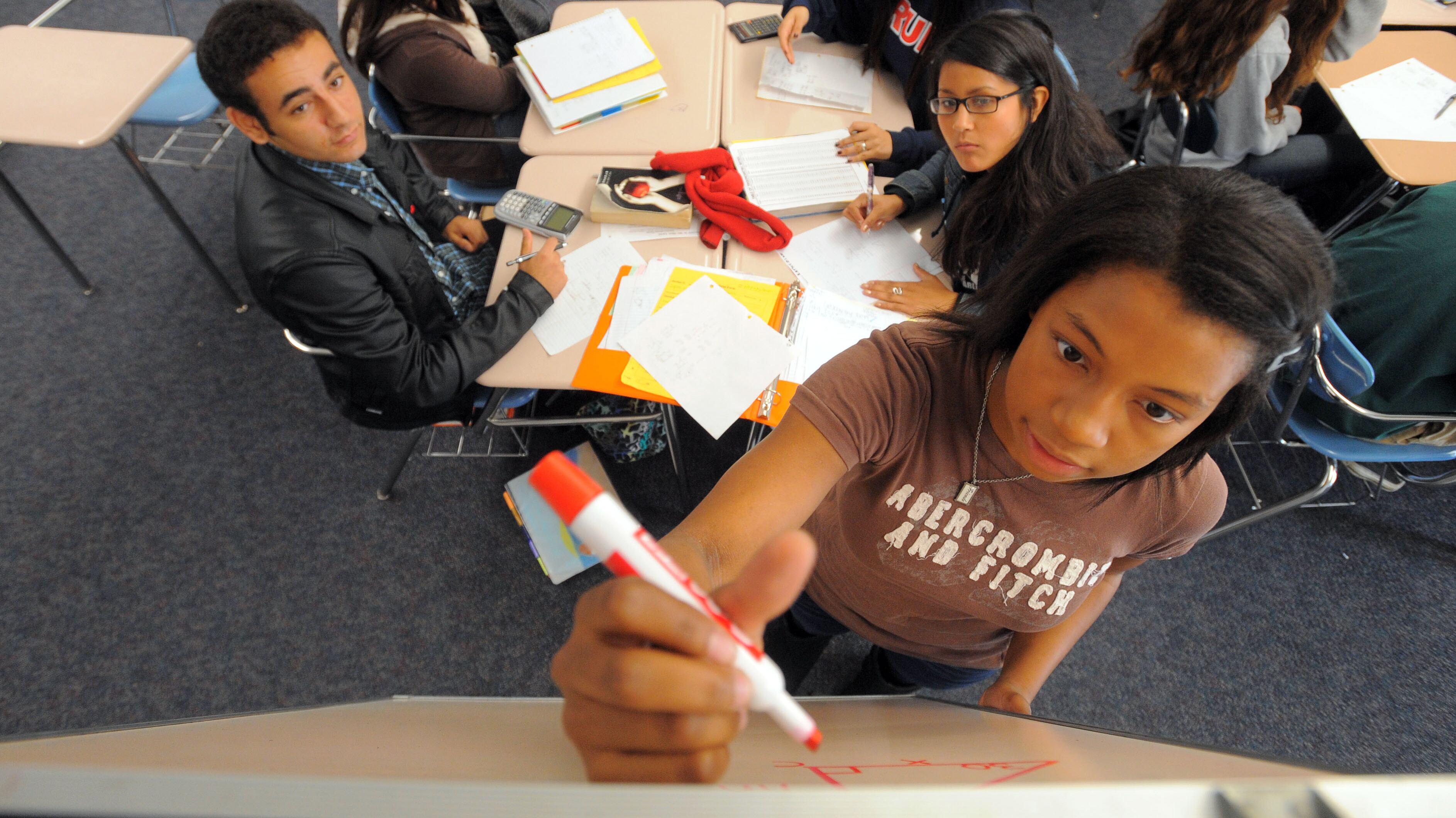 A teenage girl wearing a brown T-shirt holds a marker and writes a math problem on a whiteboard while her peers look on from a nearby desk.