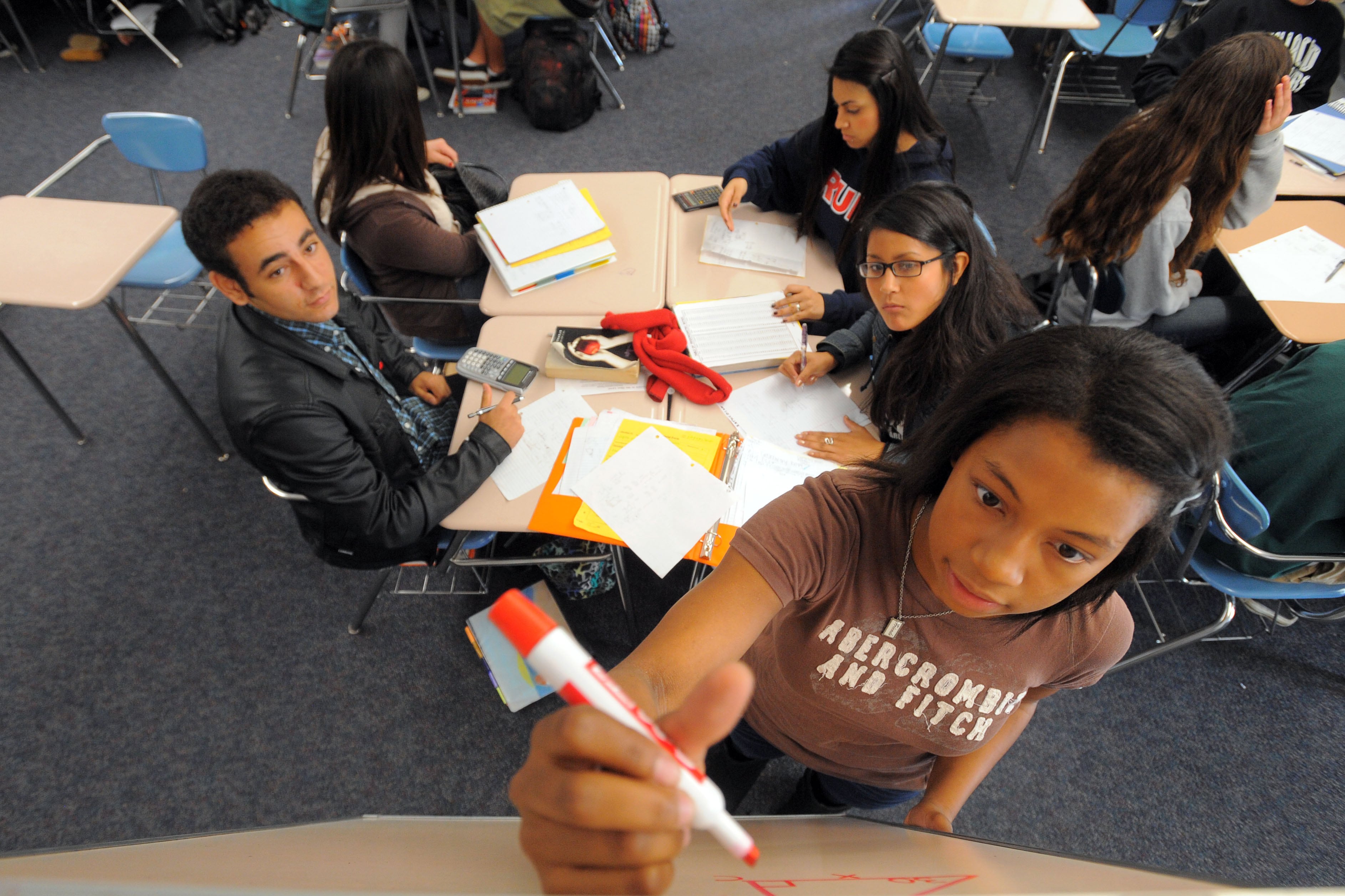 A teenage girl wearing a brown T-shirt holds a marker and writes a math problem on a whiteboard while her peers look on from a nearby desk.