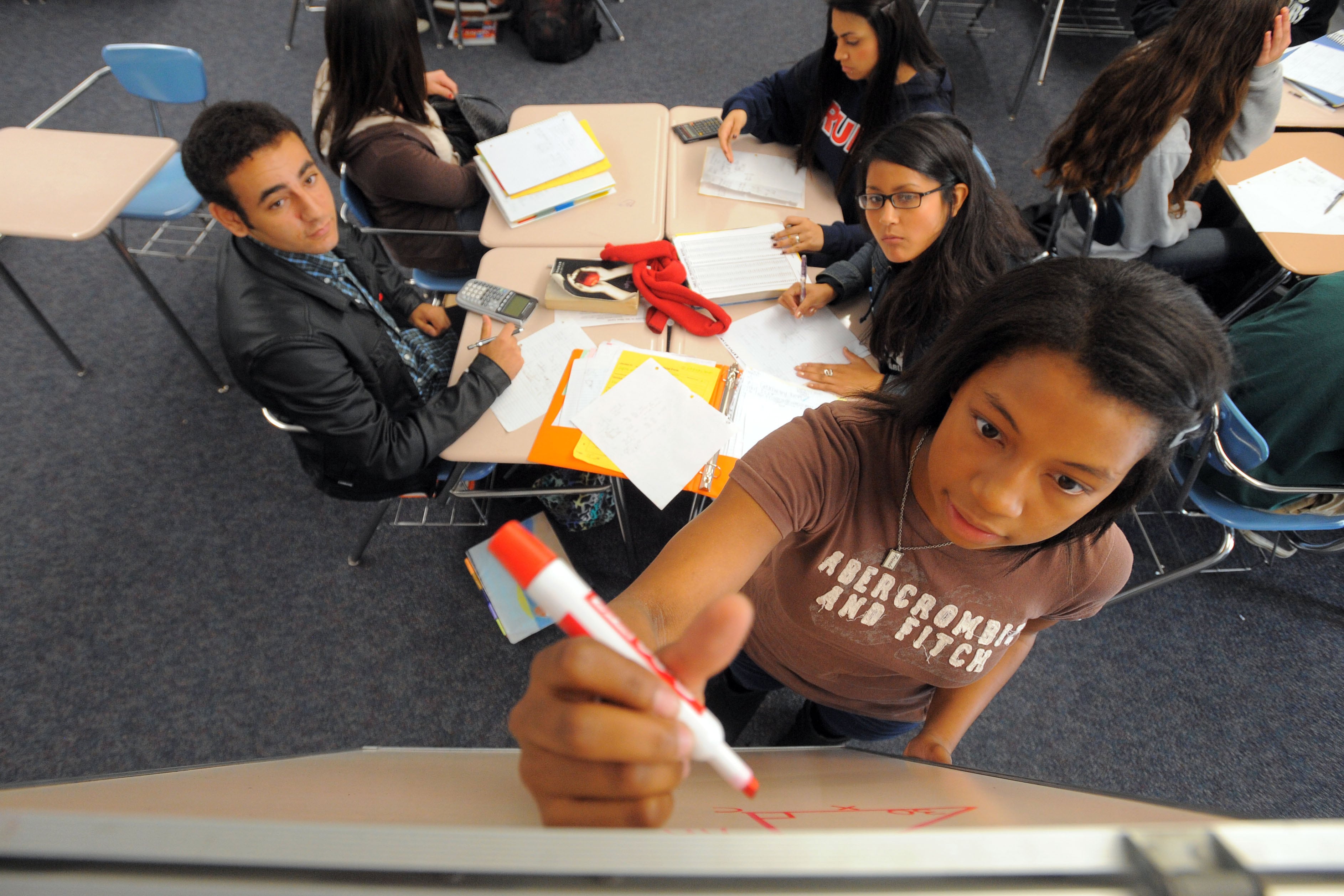 A teenage girl wearing a brown T-shirt holds a marker and writes a math problem on a whiteboard while her peers look on from a nearby desk.