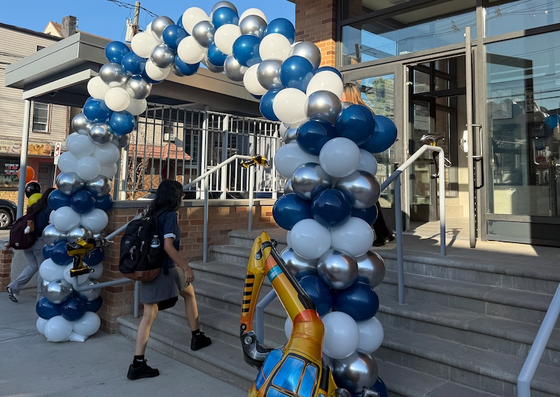 A photograph of a white and blue balloon arch in front of a large school building and one student with a backpack walks up the stairs.