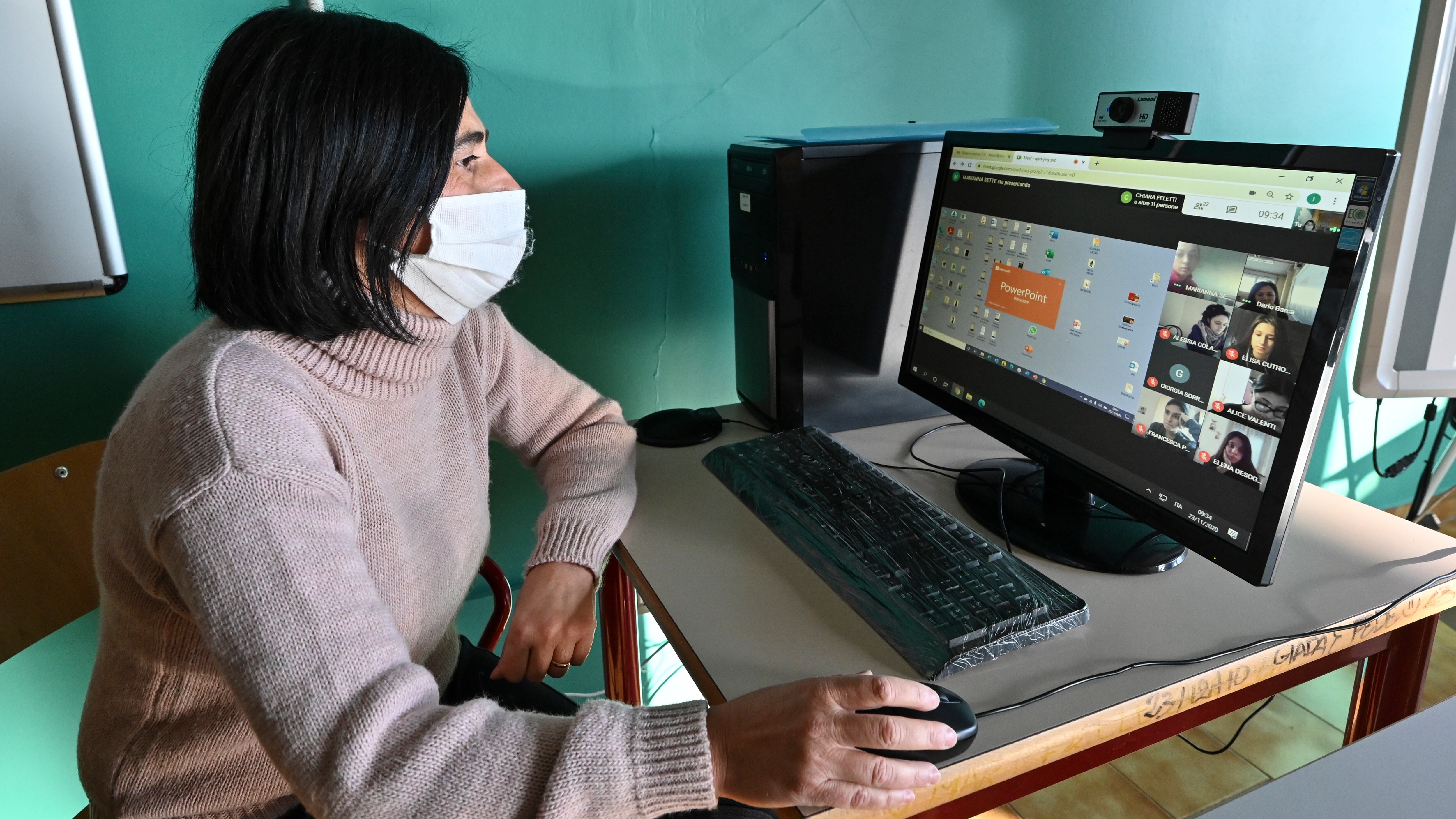 A masked female student sits at a desk and looks at a screen showing a virtual meeting.