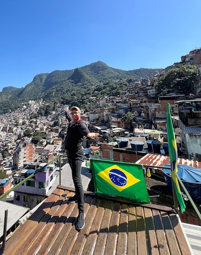 A man standing next to the Brazil flag, poses for a photo at a view point over looking a city with a blue sky.