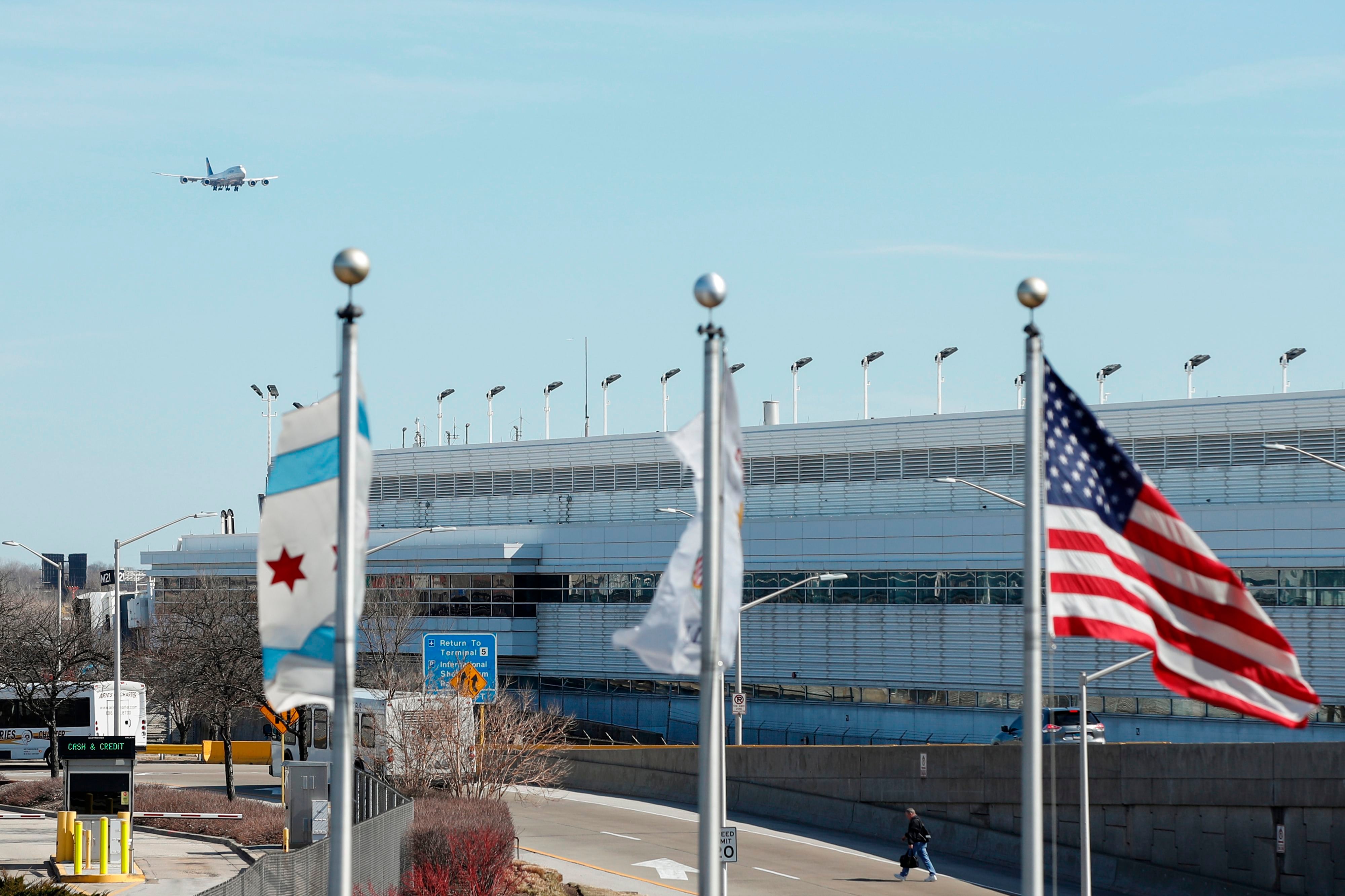 A view of Chicago O’Hare airport as a jet is flying in. The flags of Chicago, Illinois, and the United States fly in the foreground.