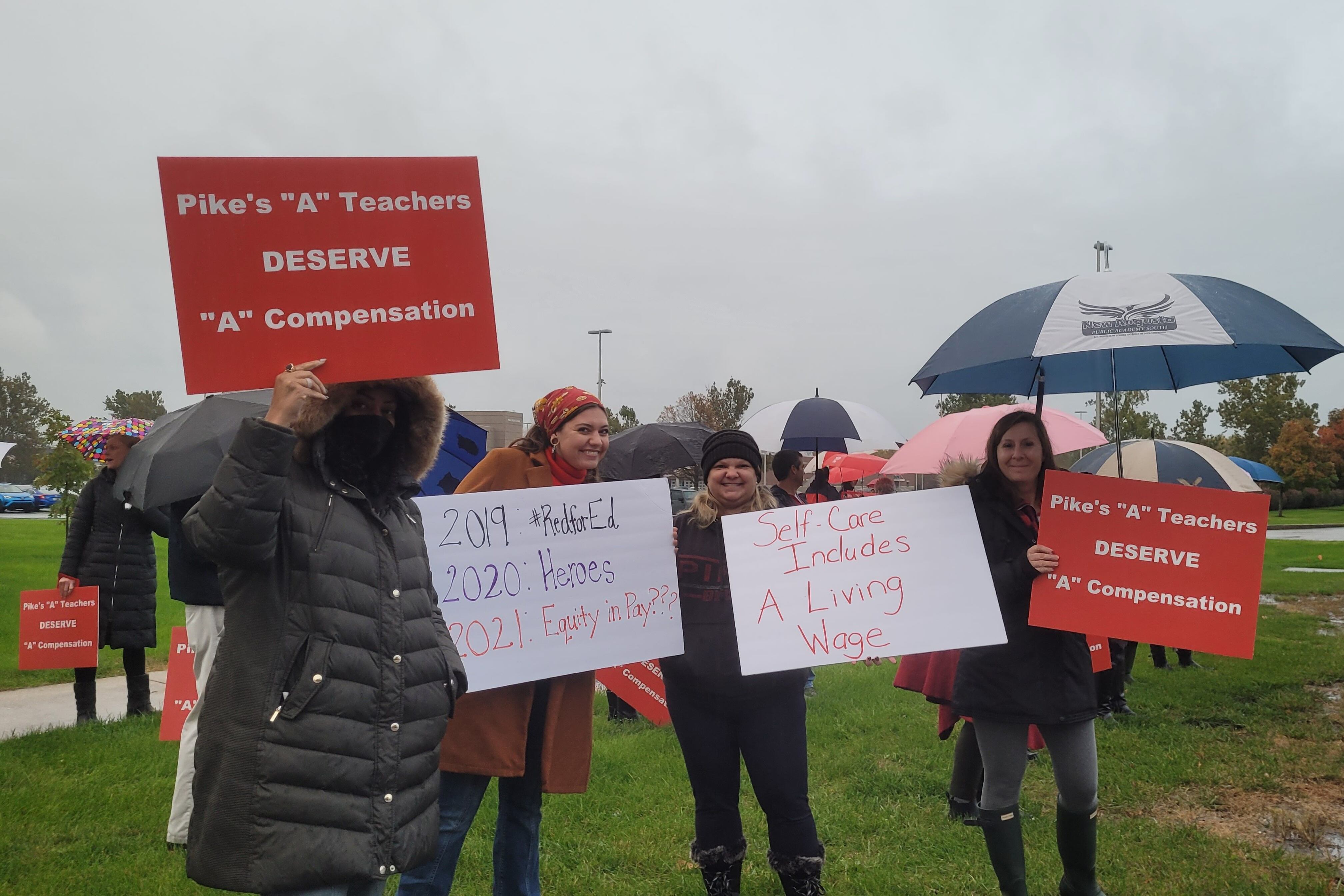 A group of people in coats and some with umbrellas stand with placards. One reads “Pike’s ‘A’ teachers deserve ‘A’ compensation” and “2019 Red for Ed, 2020 Heroes, 2021 Equity in Pay???”