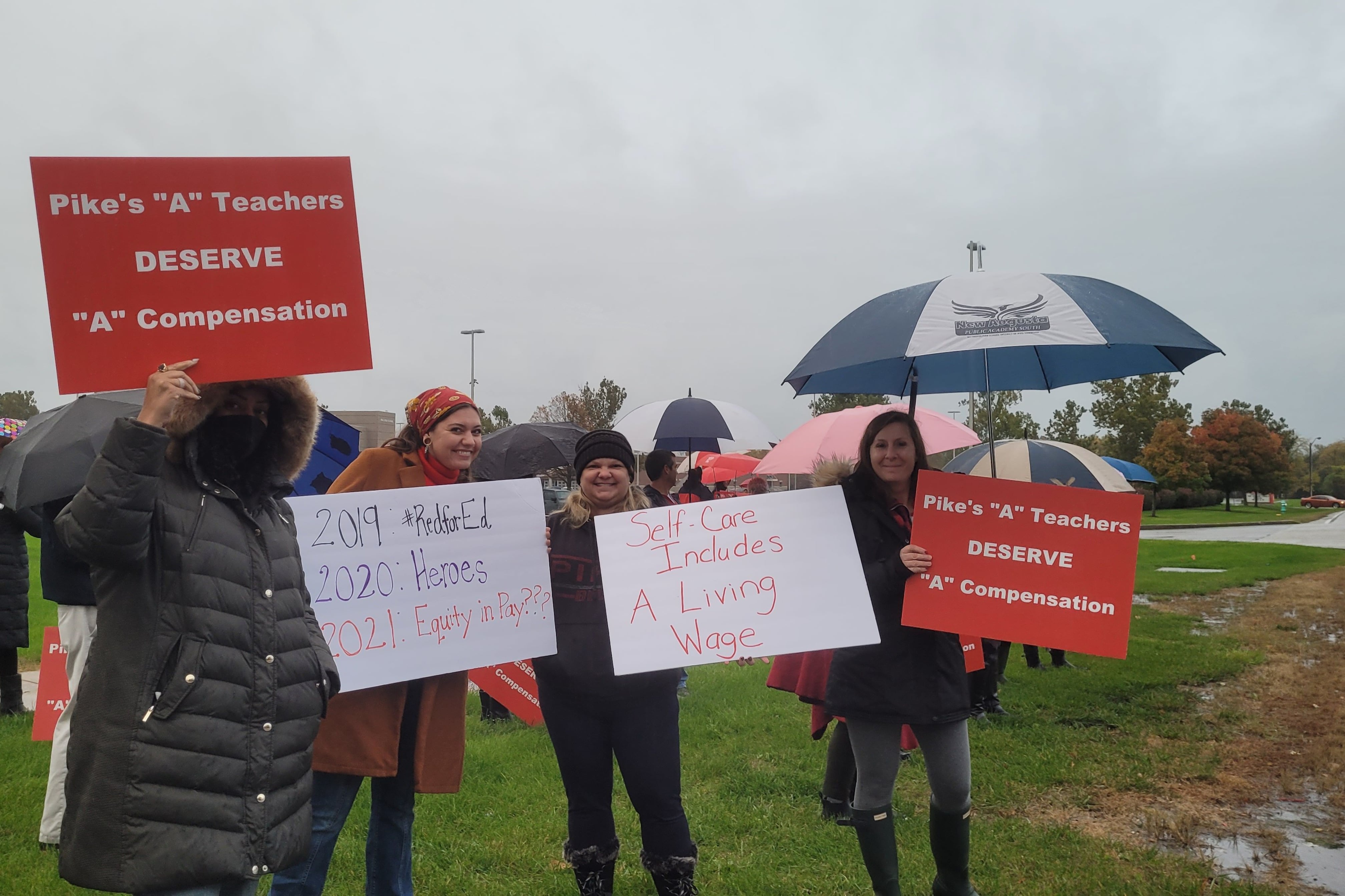 A group of people in coats and some with umbrellas stand with placards. One reads “Pike’s ‘A’ teachers deserve ‘A’ compensation” and “2019 Red for Ed, 2020 Heroes, 2021 Equity in Pay???”