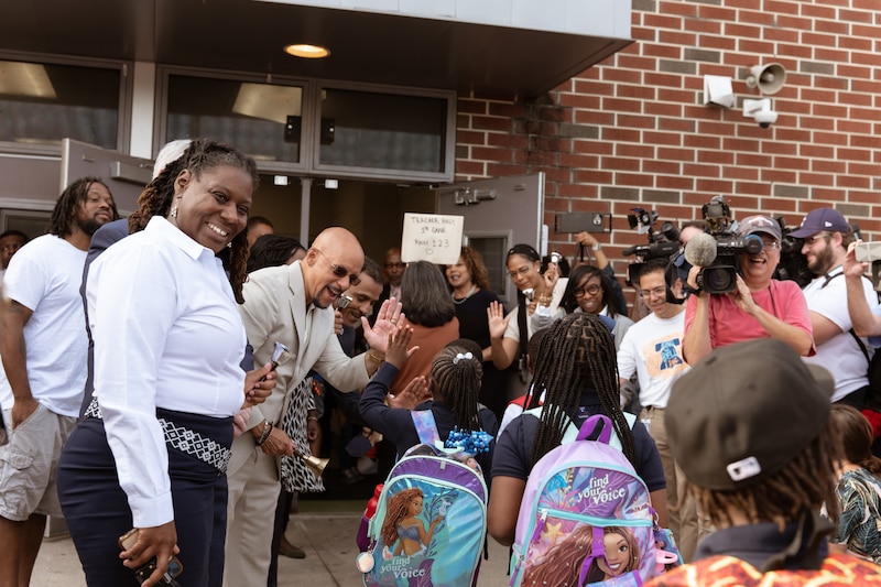 Children wearing backpacks walk into a brick building as adults and people with cameras look on.