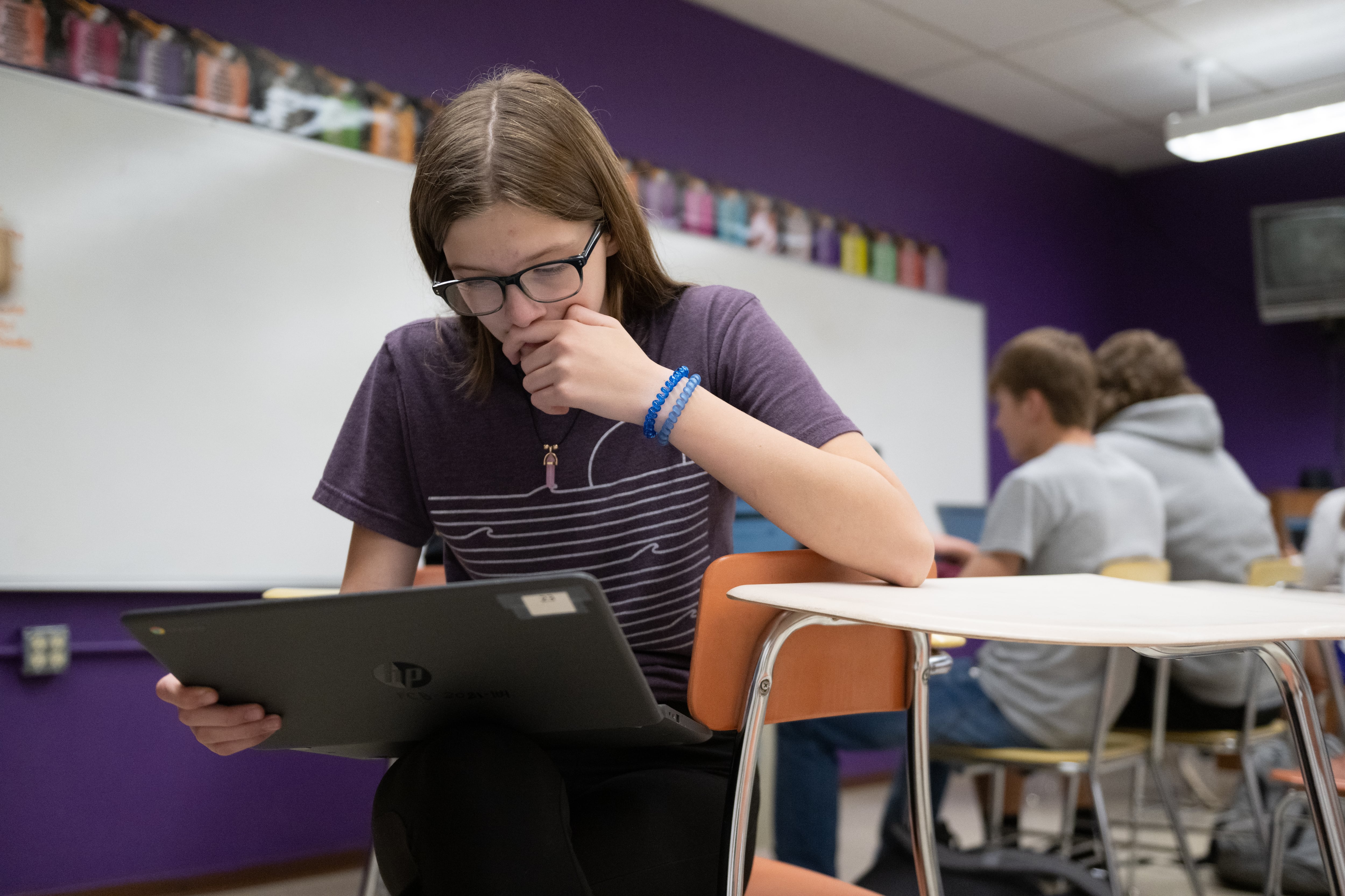 student with glasses and a purple shirt looks down at a computer screen while sitting in a classroom