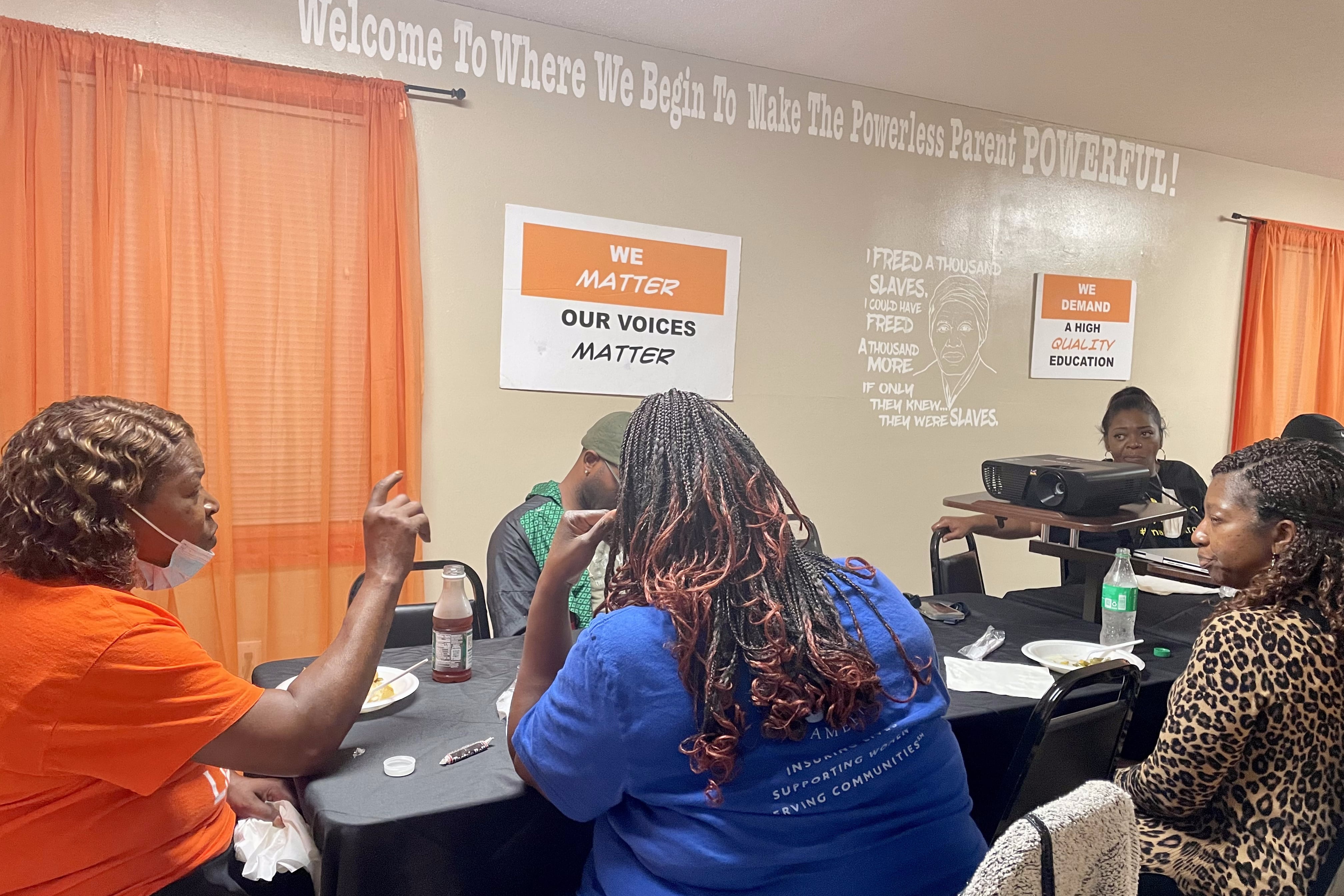 Six people sitting around a table converse in front of a wall with signs reading “We matter, our voices matter” and “Welcome to where we begin to make the powerless parent POWERFUL!”