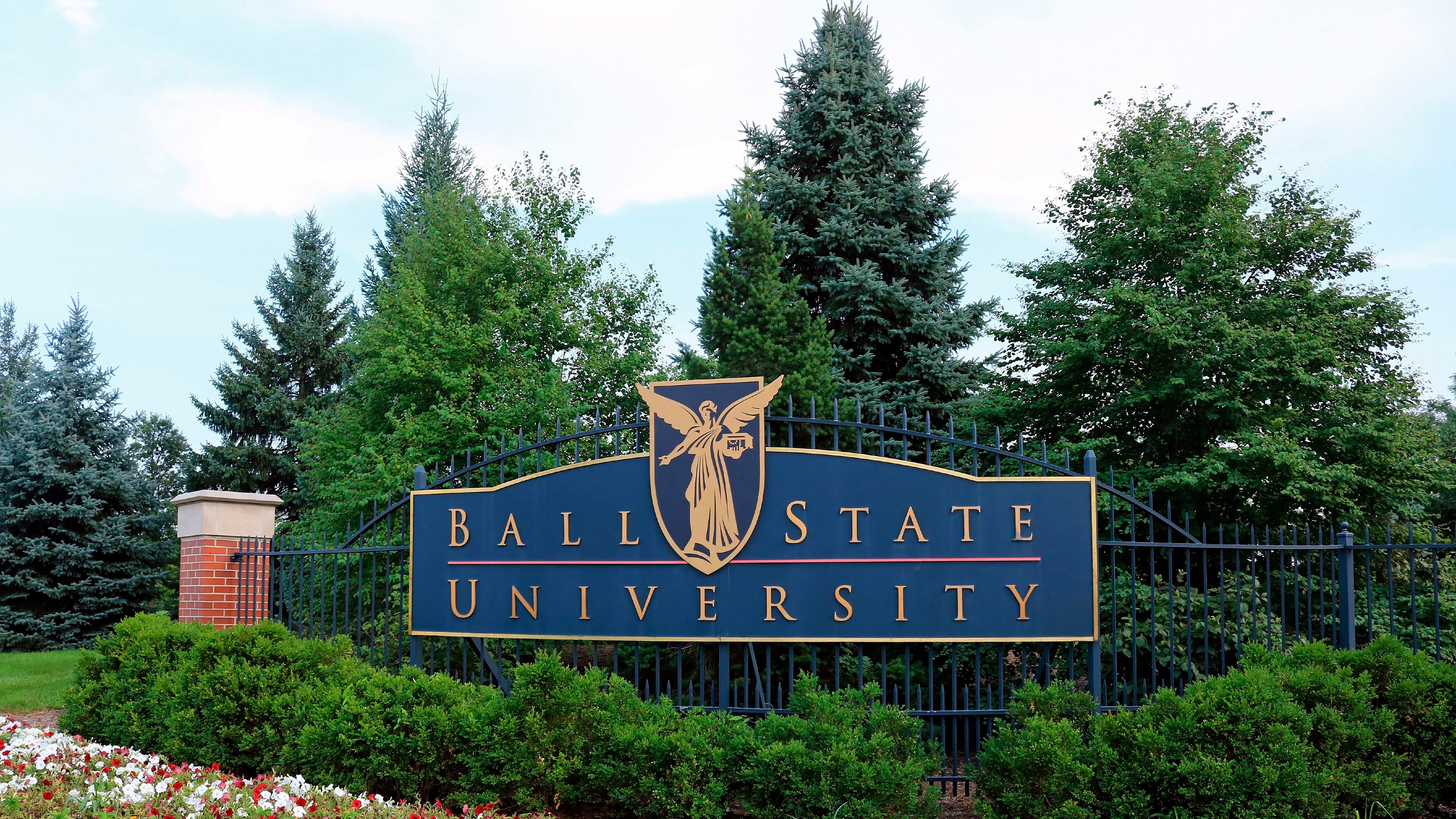 A blue sign that reads “Ball State University” hangs on a fence with coniferous trees in the background. 