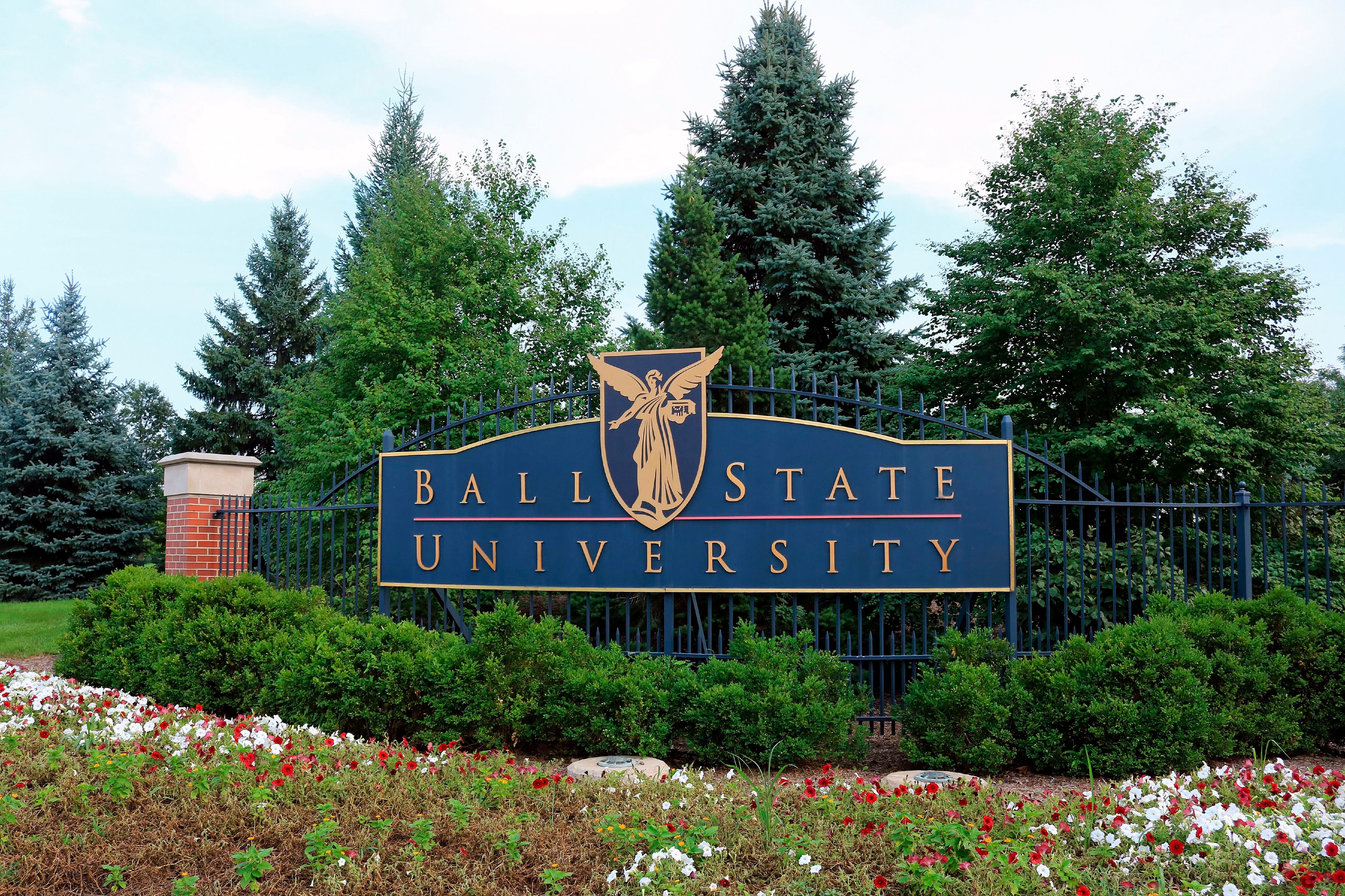 A blue sign that reads “Ball State University” hangs on a fence with coniferous trees in the background.