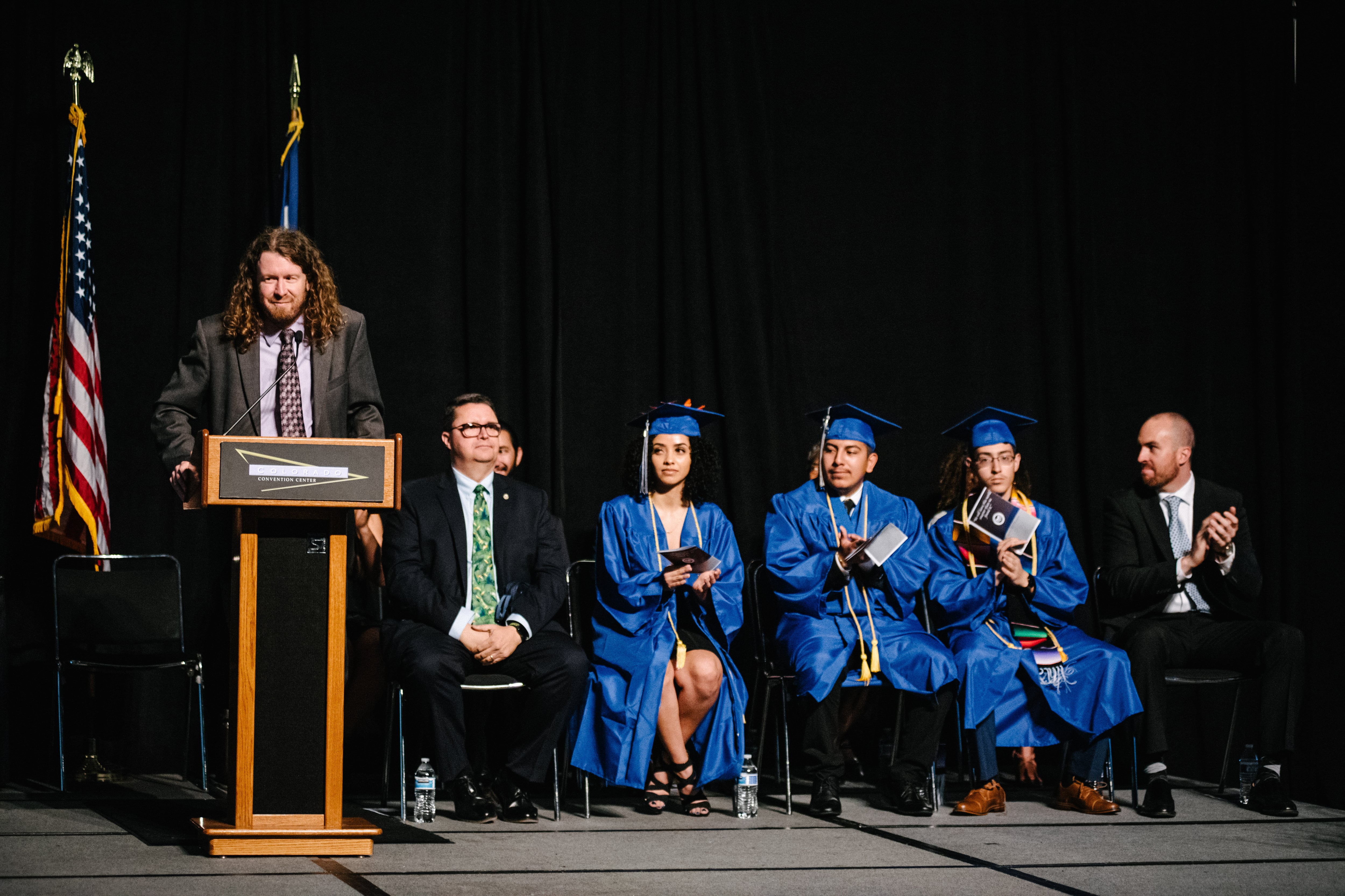 A man with shoulder-length curly red hair speaks from a lectern, dressed in a suit and tie. To his left sit two other men in suits and three teenagers in bright blue graduation caps and gowns.