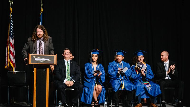 A man with shoulder-length curly red hair speaks from a lectern, dressed in a suit and tie. To his left sit two other men in suits and three teenagers in bright blue graduation caps and gowns. 