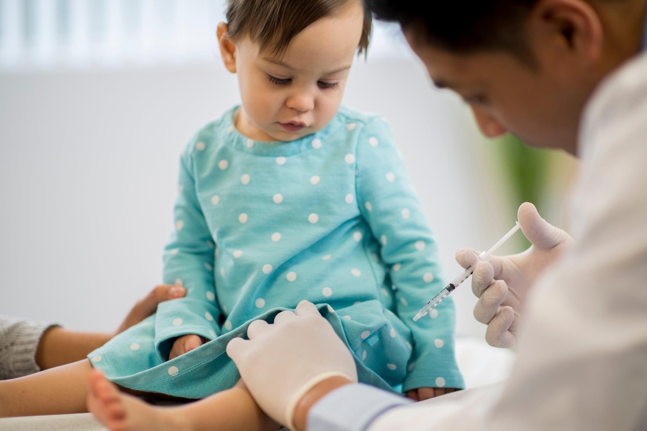 A baby sits up wearing a blue dress while a doctor gives her a vaccine.