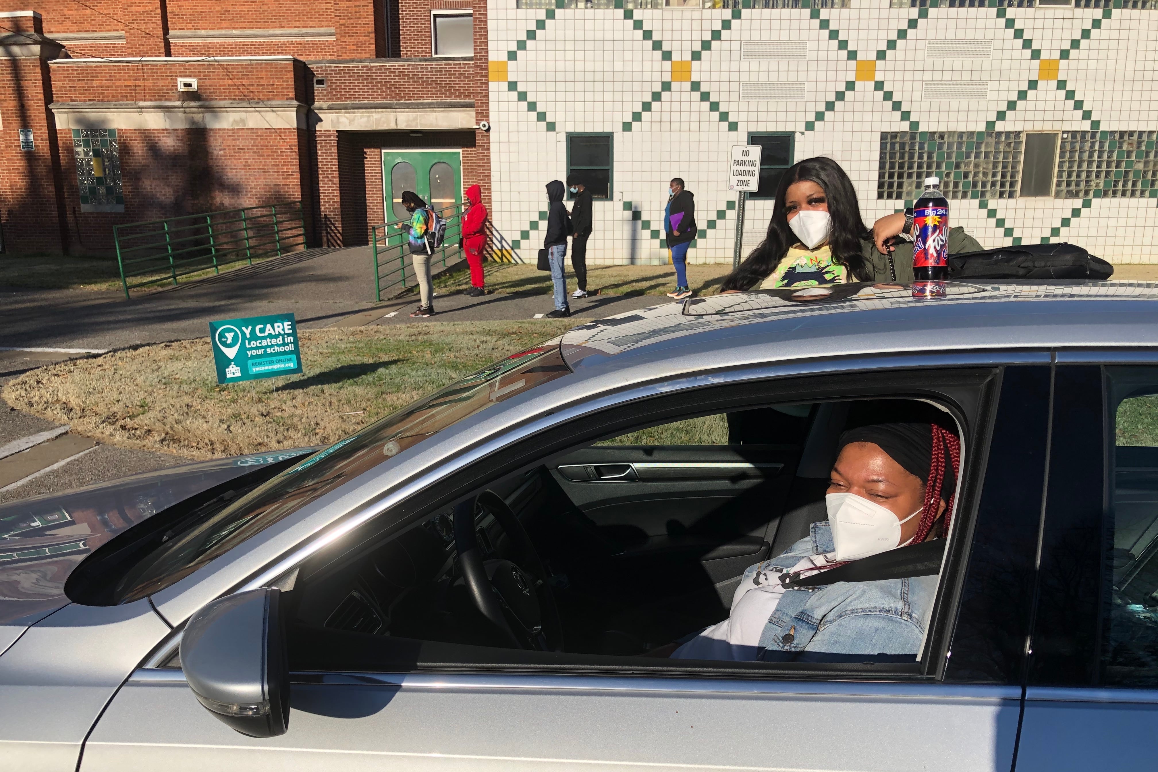 A masked parent in the driver’s seat of a car and her masked daughter exiting the car in front of the school, as students stand apart in line to enter the building.