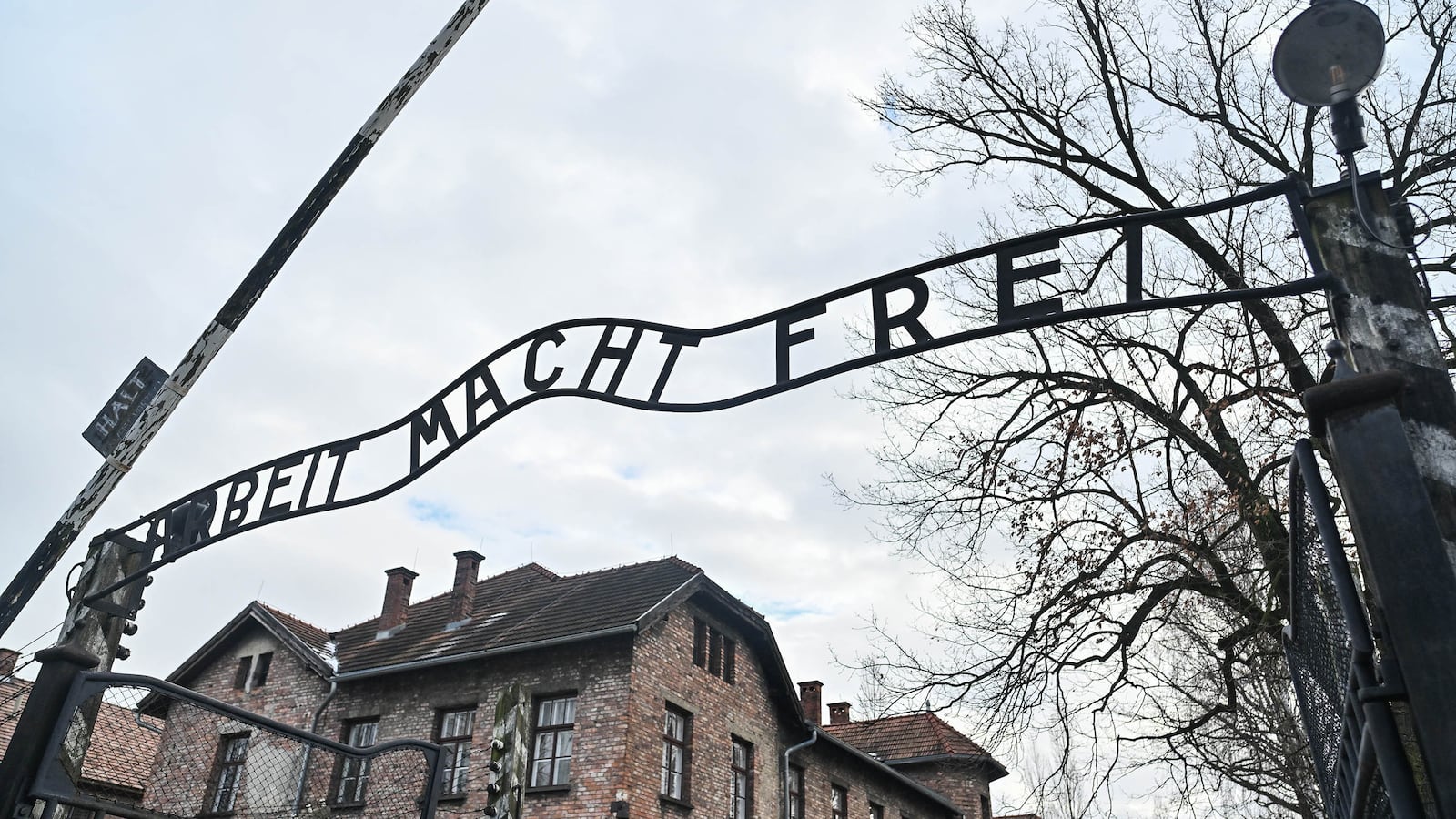 A metal gate with a sign that reads 'Arbeit Macht Frei' (Work Sets You Free) stands above a tan brick building in the background with trees and a cloudy sky.