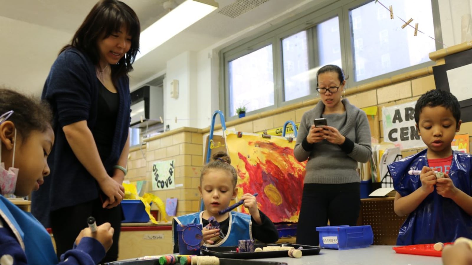 Yvette Ho, right, taps out a request to NYCHA to fix a leaky roof at CPC Jacob Riis Child Care Center. Meanwhile, a student shows her art project to Mary Cheng, who oversees early childhood programs for the Chinese-American Planning Council, a nonprofit that runs the daycare.
