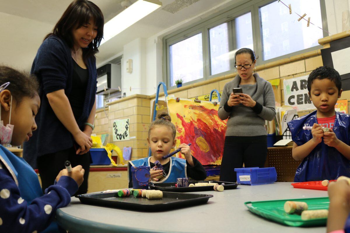 Yvette Ho, right, taps out a request to NYCHA to fix a leaky roof at CPC Jacob Riis Child Care Center. Meanwhile, a student shows her art project to Mary Cheng, who oversees early childhood programs for the Chinese-American Planning Council, a nonprofit that runs the daycare.