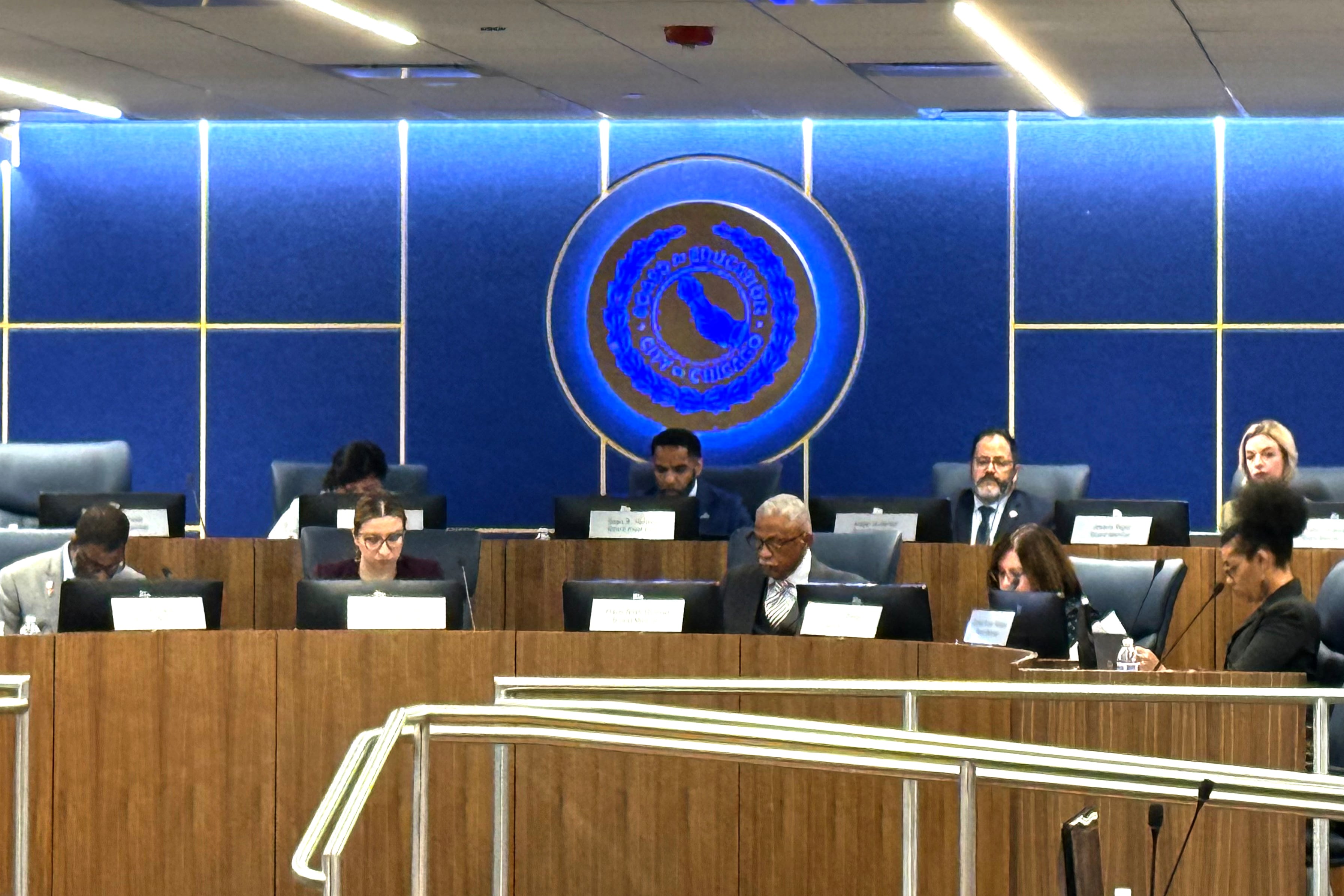 Men and women set at long desks in front of a seal.