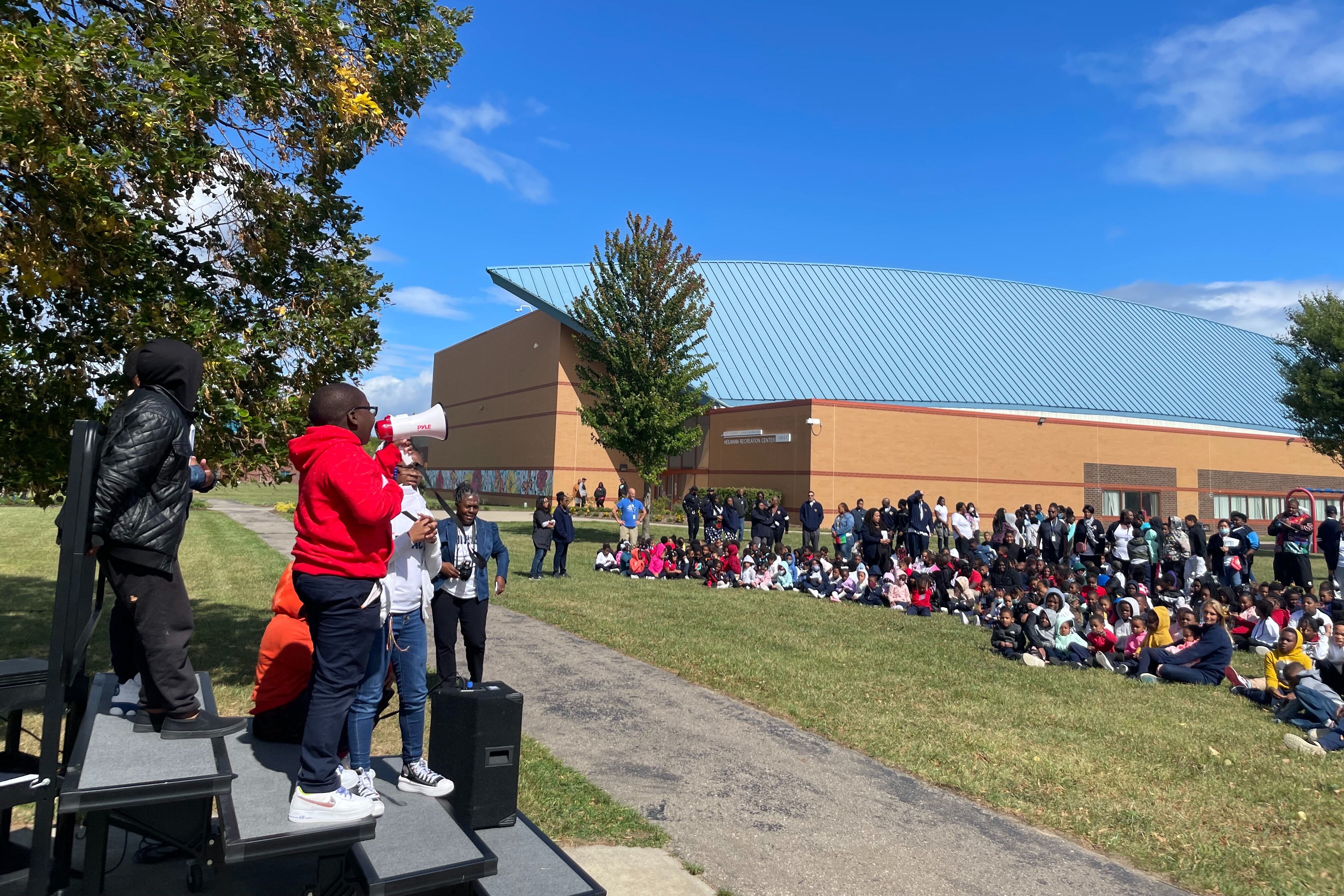 A child dressed in a red hoodie is holding a megaphone and speaking in front of a crowd of adults and young people.