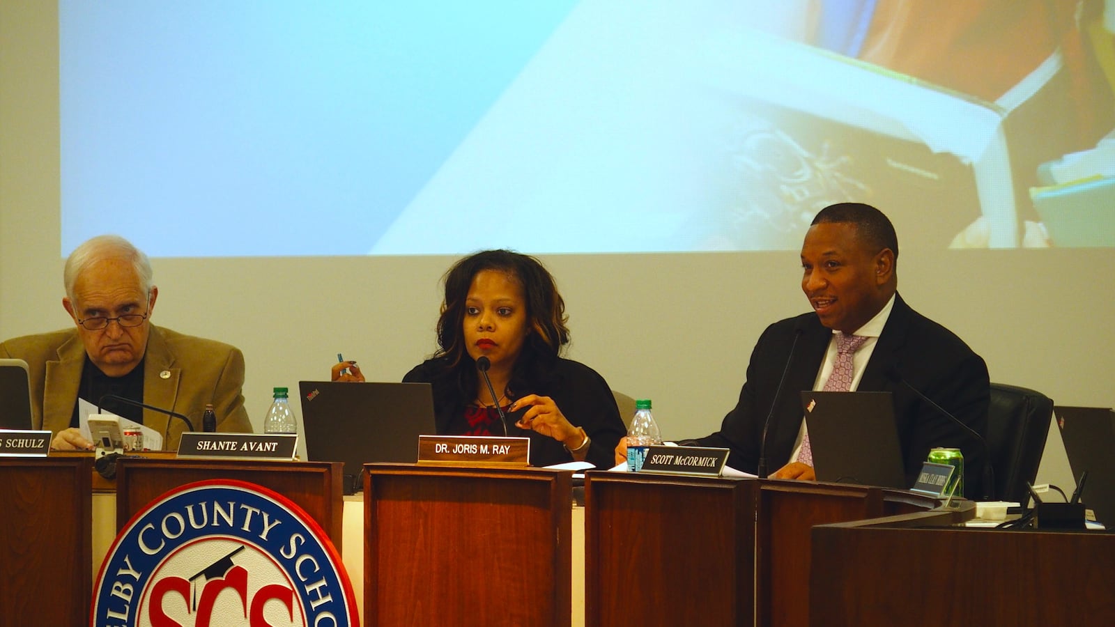 A woman, sitting next to two men at a podium, speaks into a microphone.