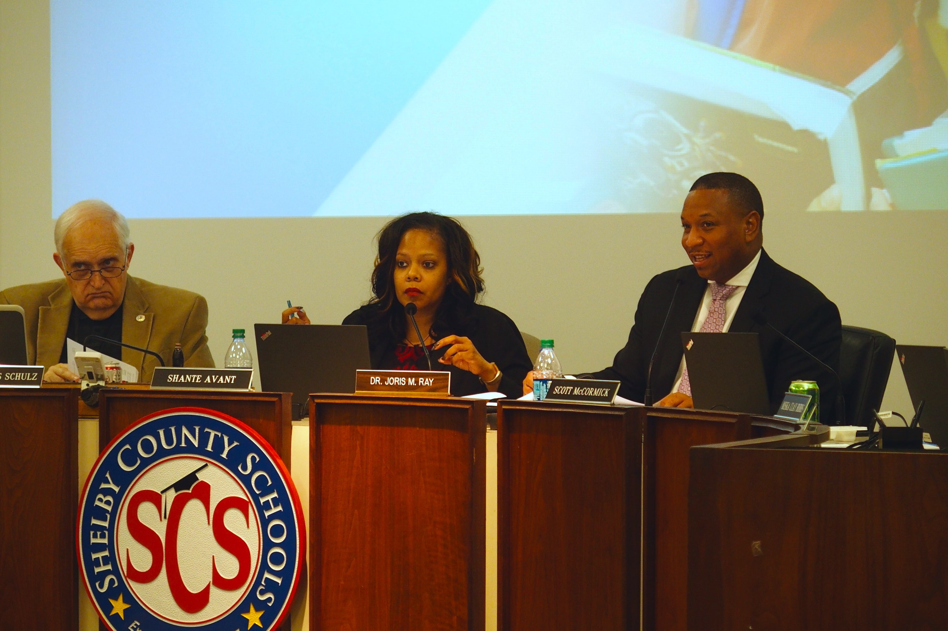 A woman, sitting next to two men at a podium, speaks into a microphone.