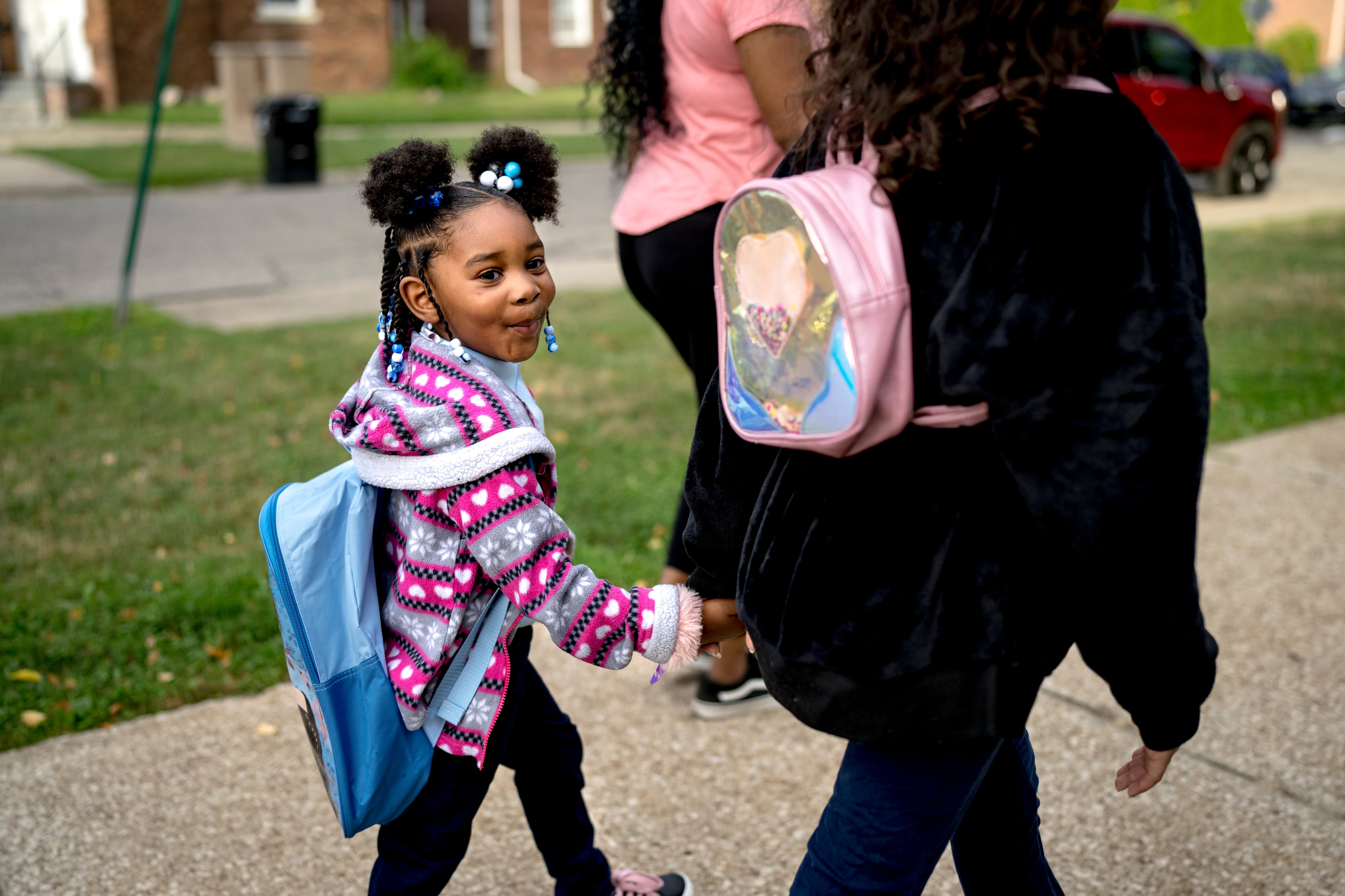 A young girl wearing a sweater and a backpack holds the hand of an adult while walking down the street.