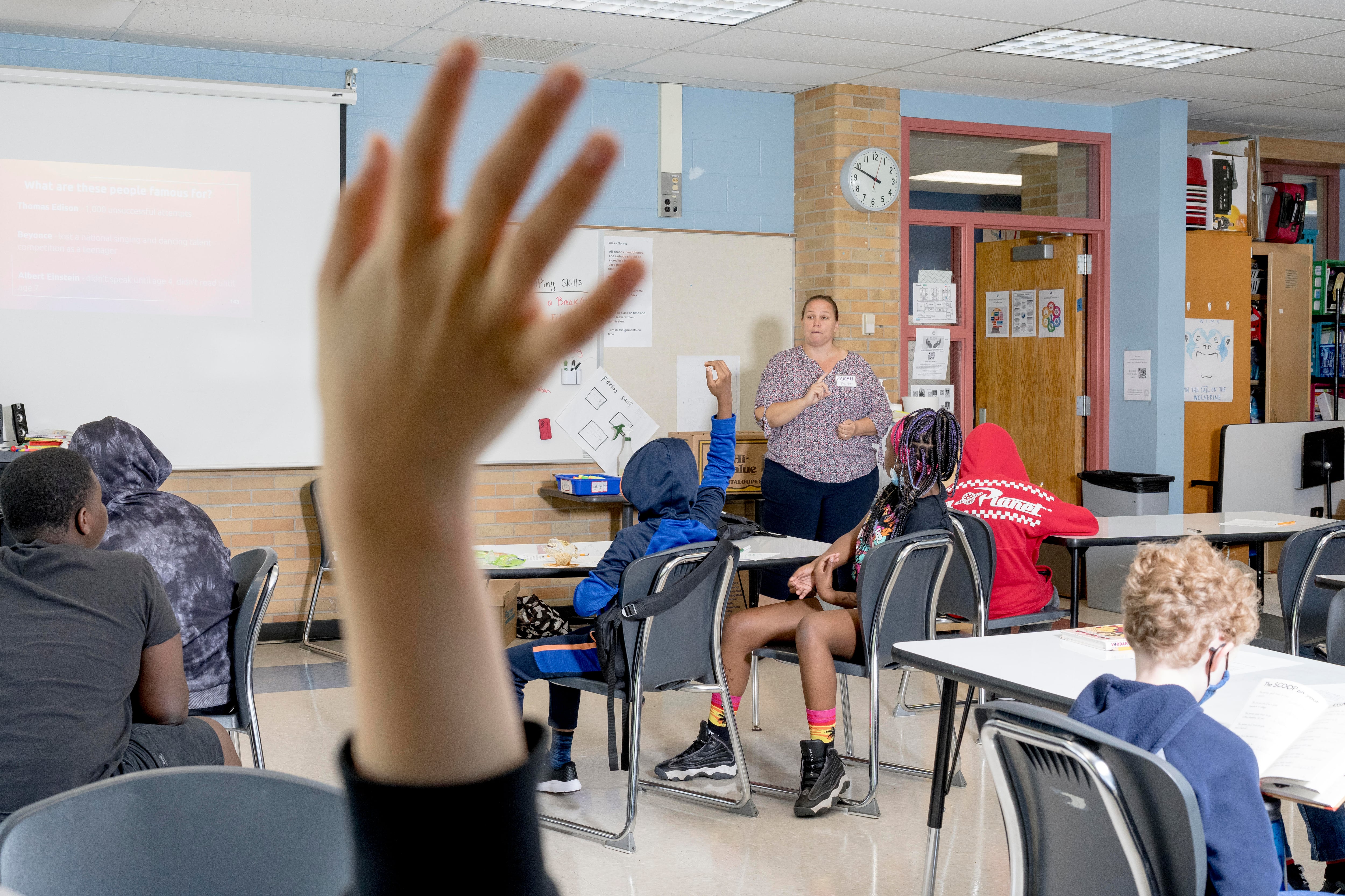 Several students sit at desks in a large classroom. Two of them raise their hands while a teacher stands at the front of the room.