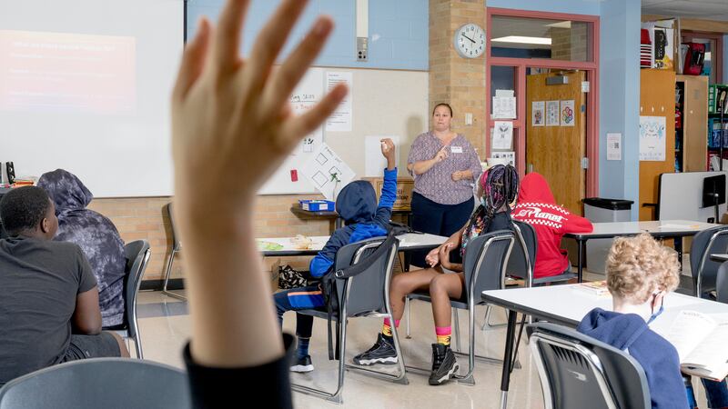 Several students sit at desks in a large classroom. Two of them raise their hands while a teacher stands at the front of the room.