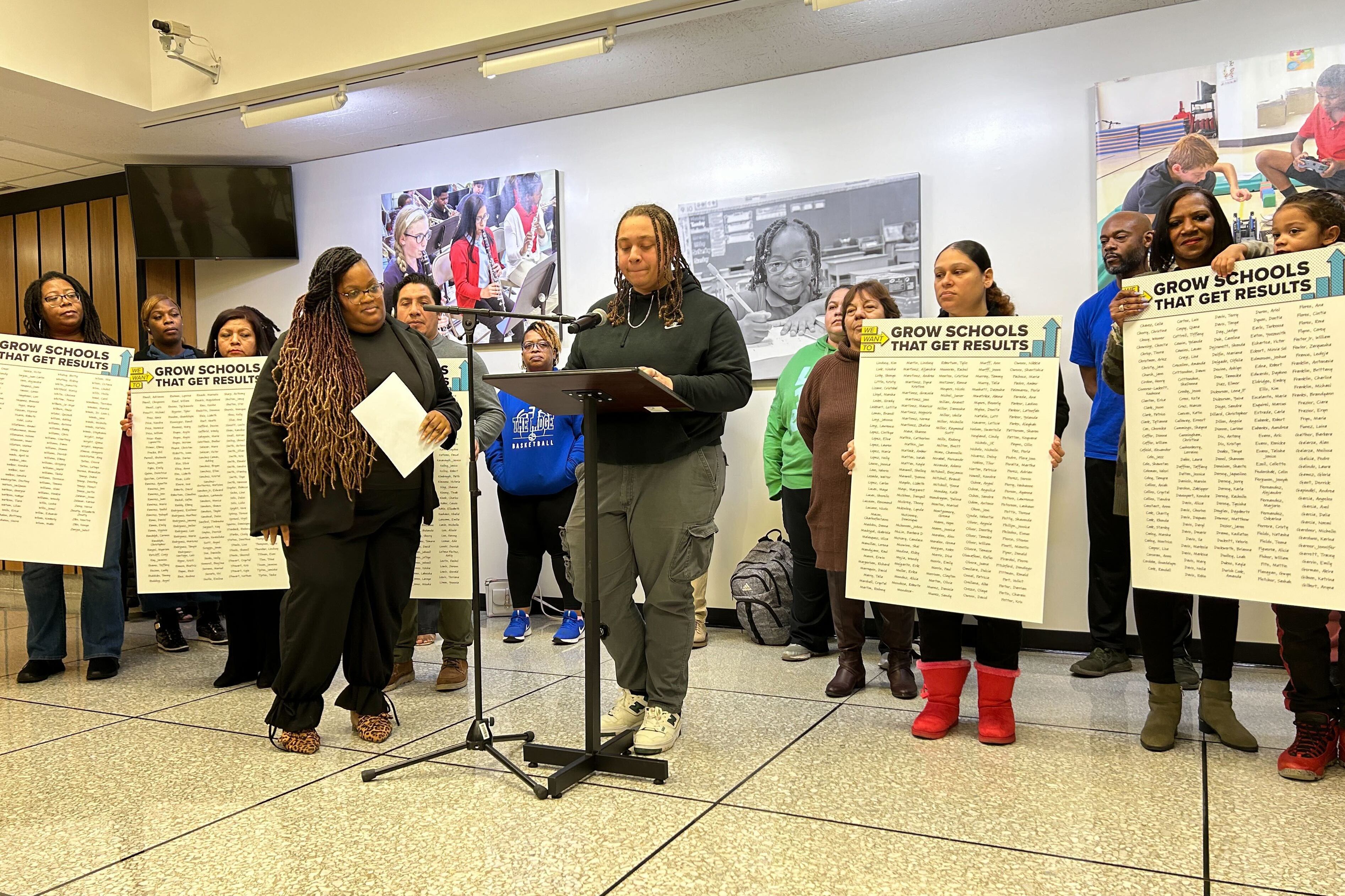 A student with long braids and wearing a black sweater stands at a podium while a group of people stand in the background holding up signs.