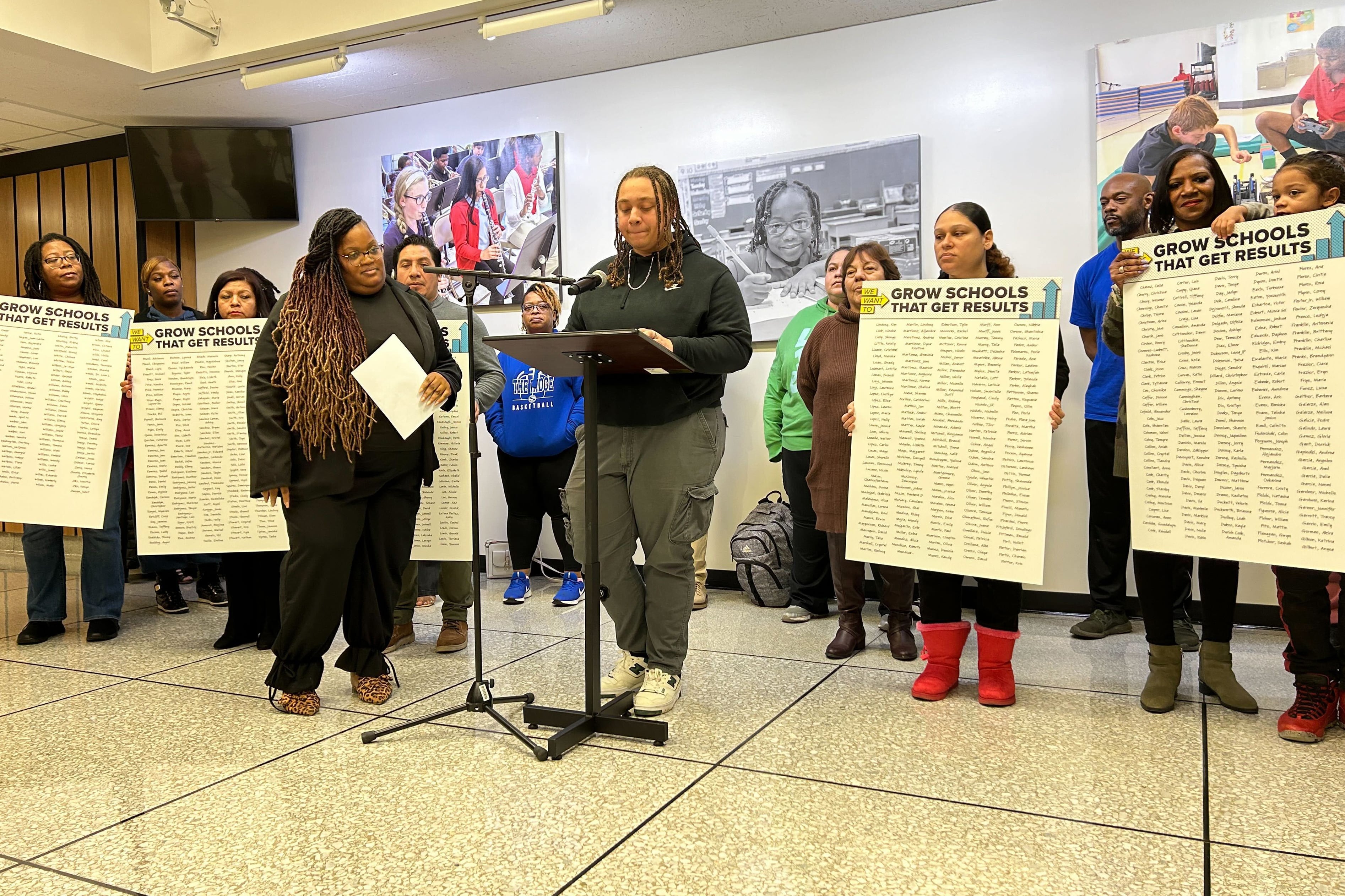 A student with long braids and wearing a black sweater stands at a podium while a group of people stand in the background holding up signs.