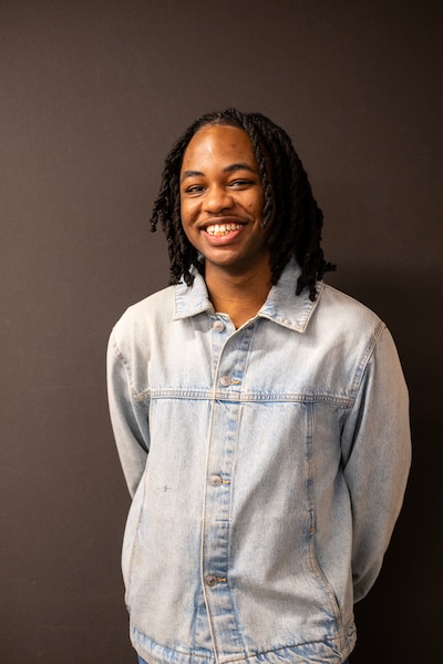 A photograph of a Black high school senior with short dark twists and wearing a light dress shirt poses with a big smile against a brown background.