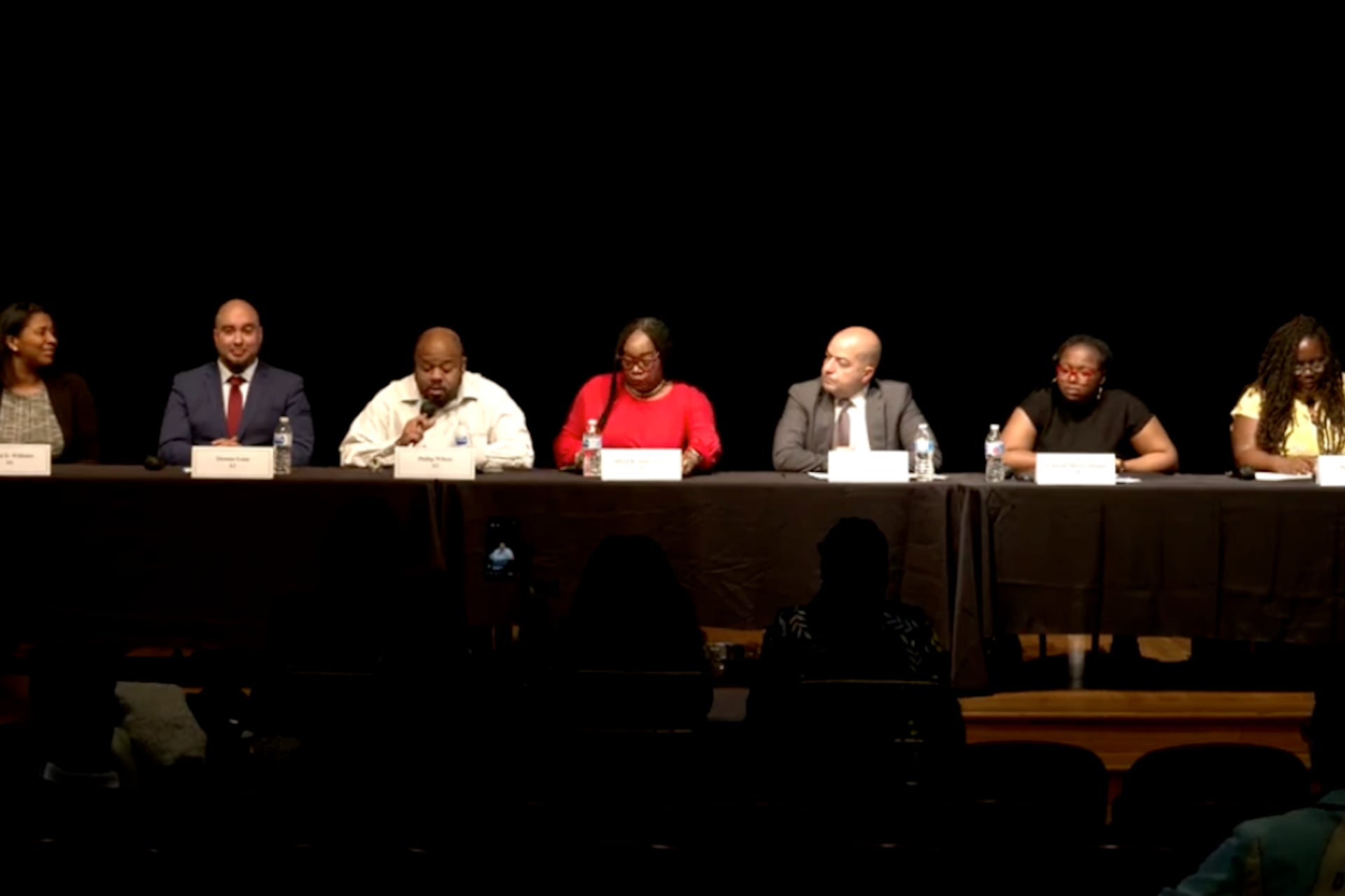 From left, Crystal Williams, Thomas Luna, Phillip Wilson, Allison K. James-Frison, Daniel Gonzalez, A’Dorian Murray-Thomas, and Maggie Freeman sit at a table on stage with a black background.
