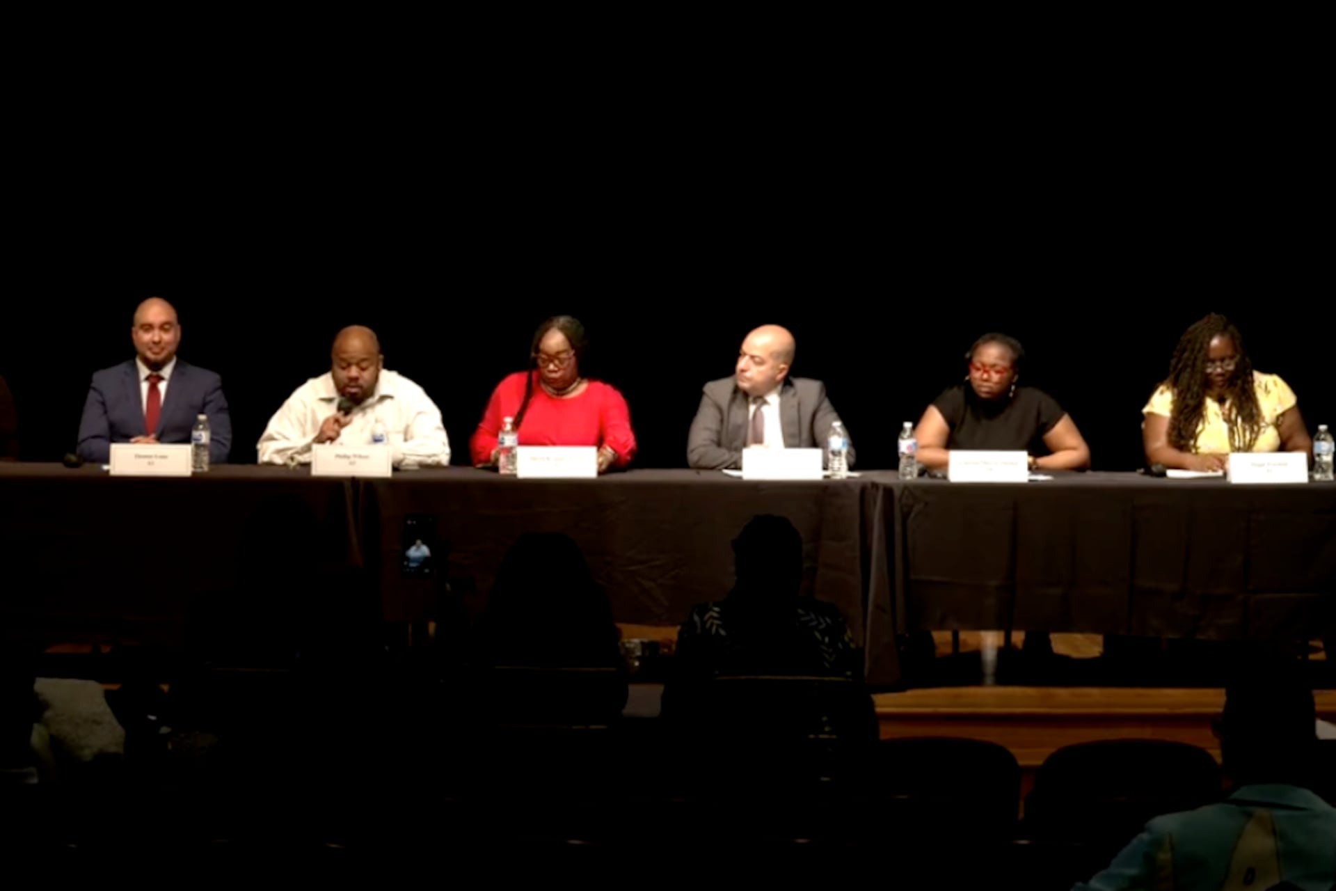 From left, Crystal Williams, Thomas Luna, Phillip Wilson, Allison K. James-Frison, Daniel Gonzalez, A’Dorian Murray-Thomas, and Maggie Freeman sit at a table on stage with a black background.
