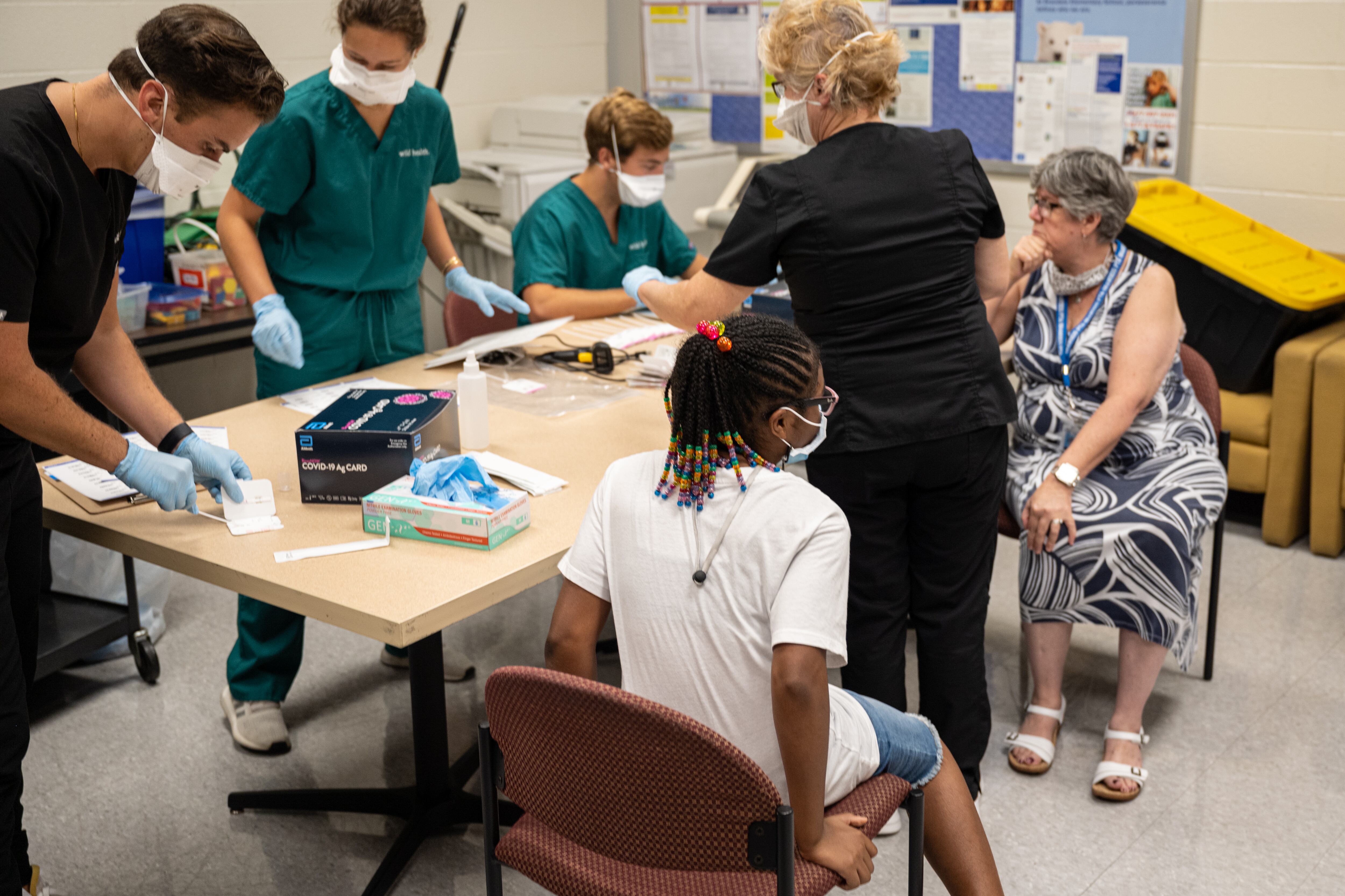 People with masks on gather around a table with needles. A child and elderly woman sit at the edge of the table waiting for a COVID-19 test.