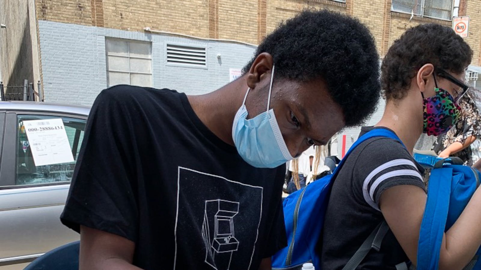 A high school student with short dark hair and wearing a face mask works at a desk in a classroom.