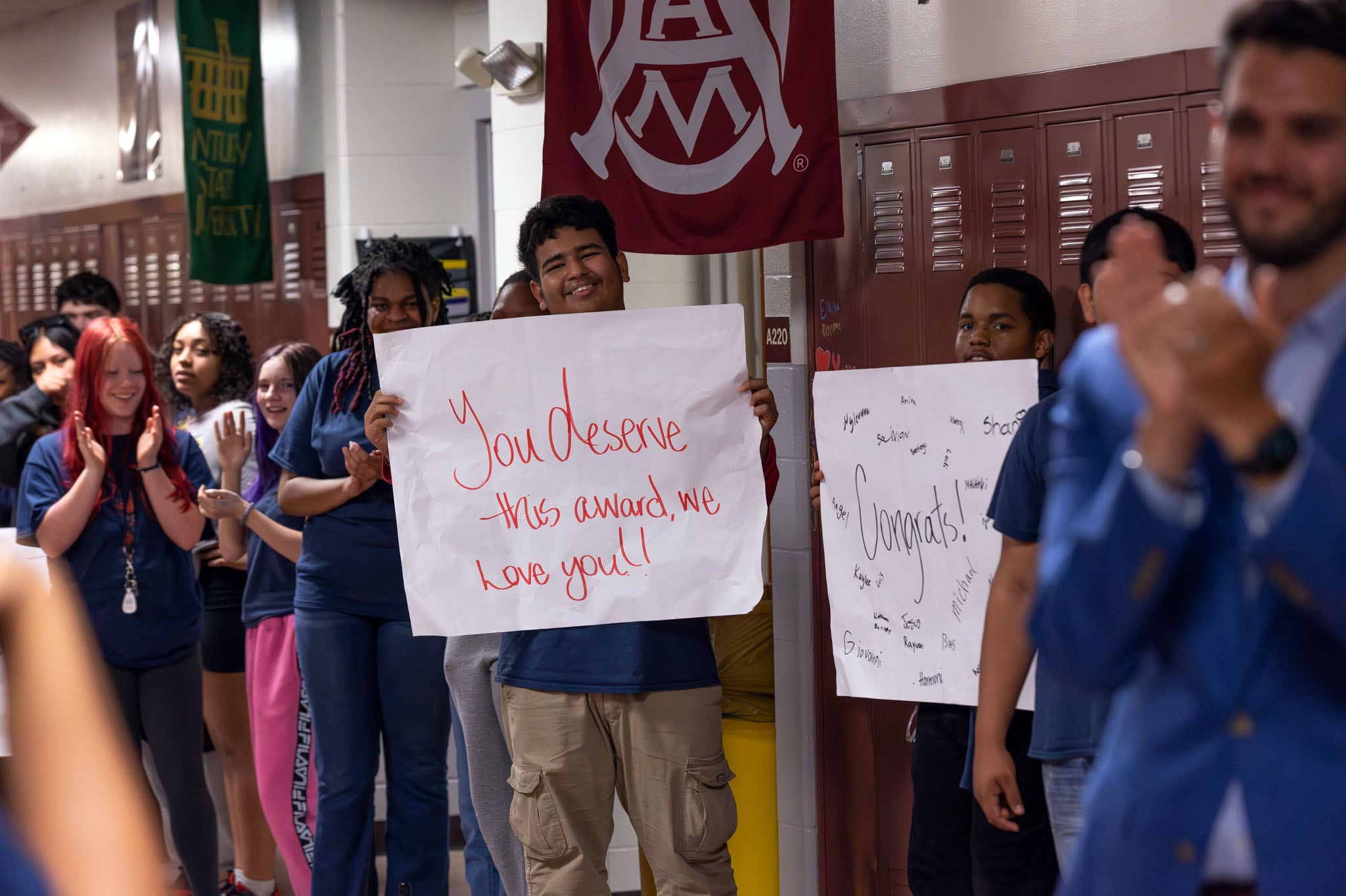 A group of adults and students line a hallway with one person holding up a large white sign that reads "You deserve this award, we love you."