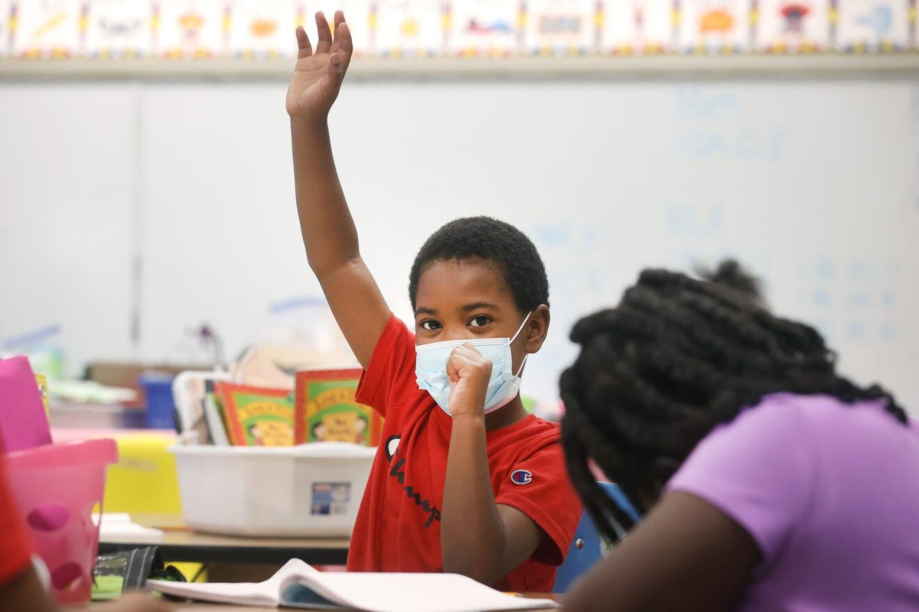 A student wearing a red shirt and protective mask raises his hand in an elementary school classroom.