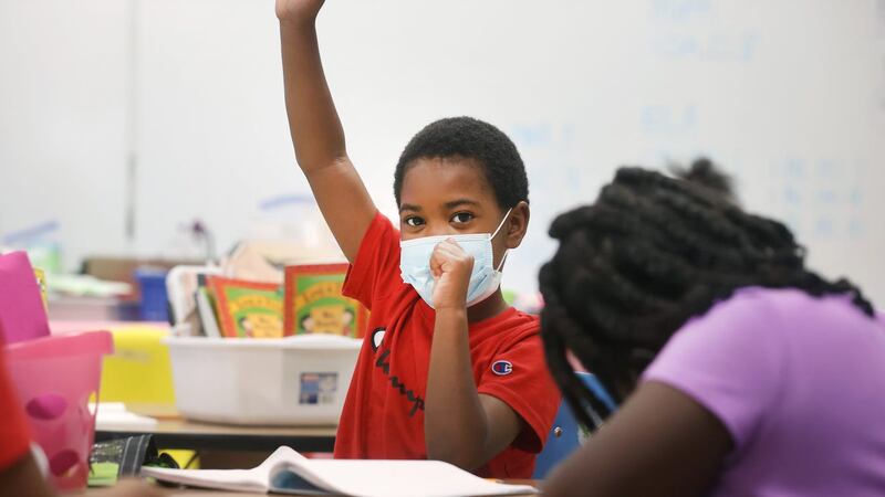 A student wearing a red shirt and protective mask raises his hand in an elementary school classroom.